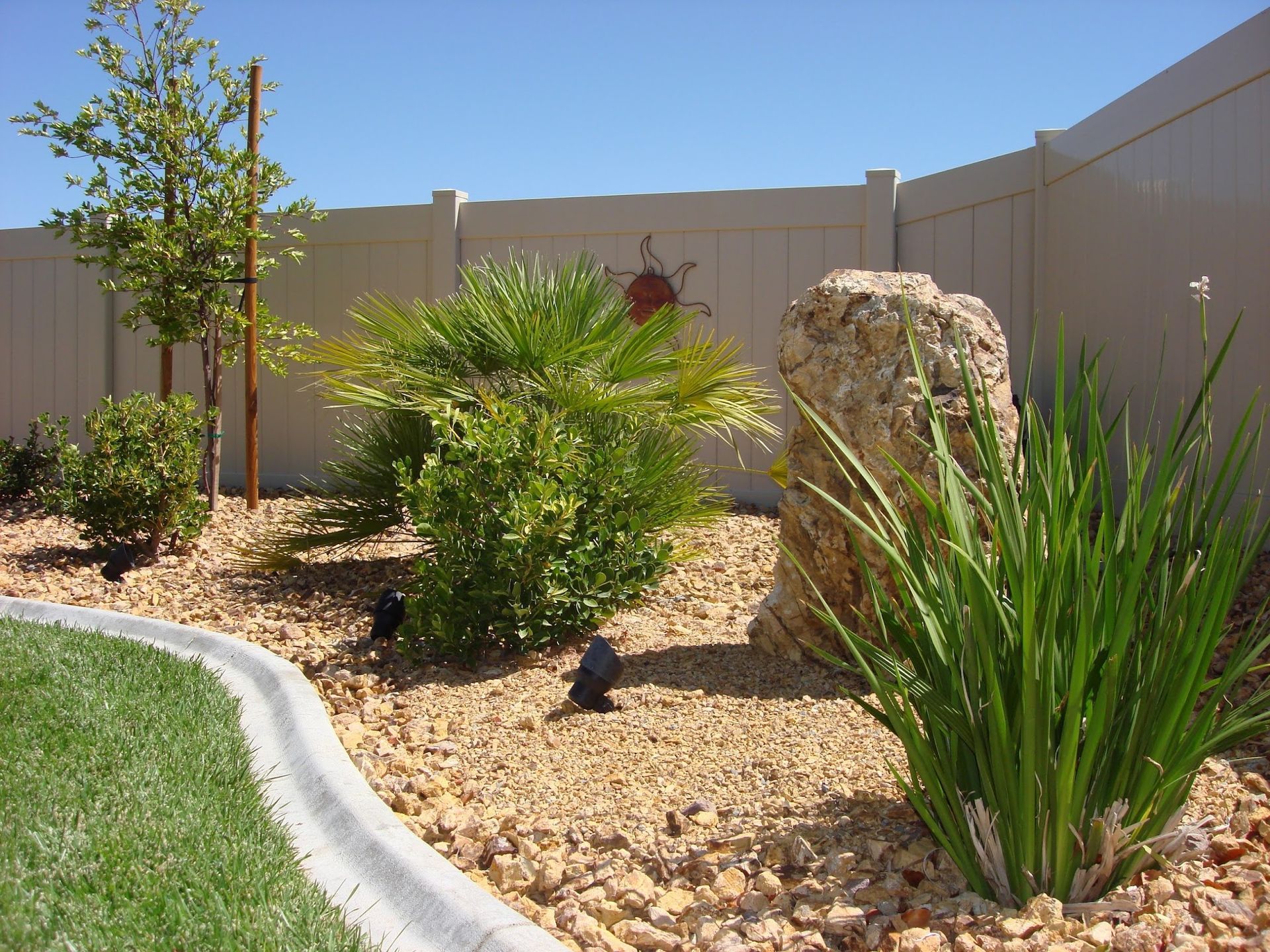 Backyard garden with beige gravel, plants, a large rock, and a beige fence.