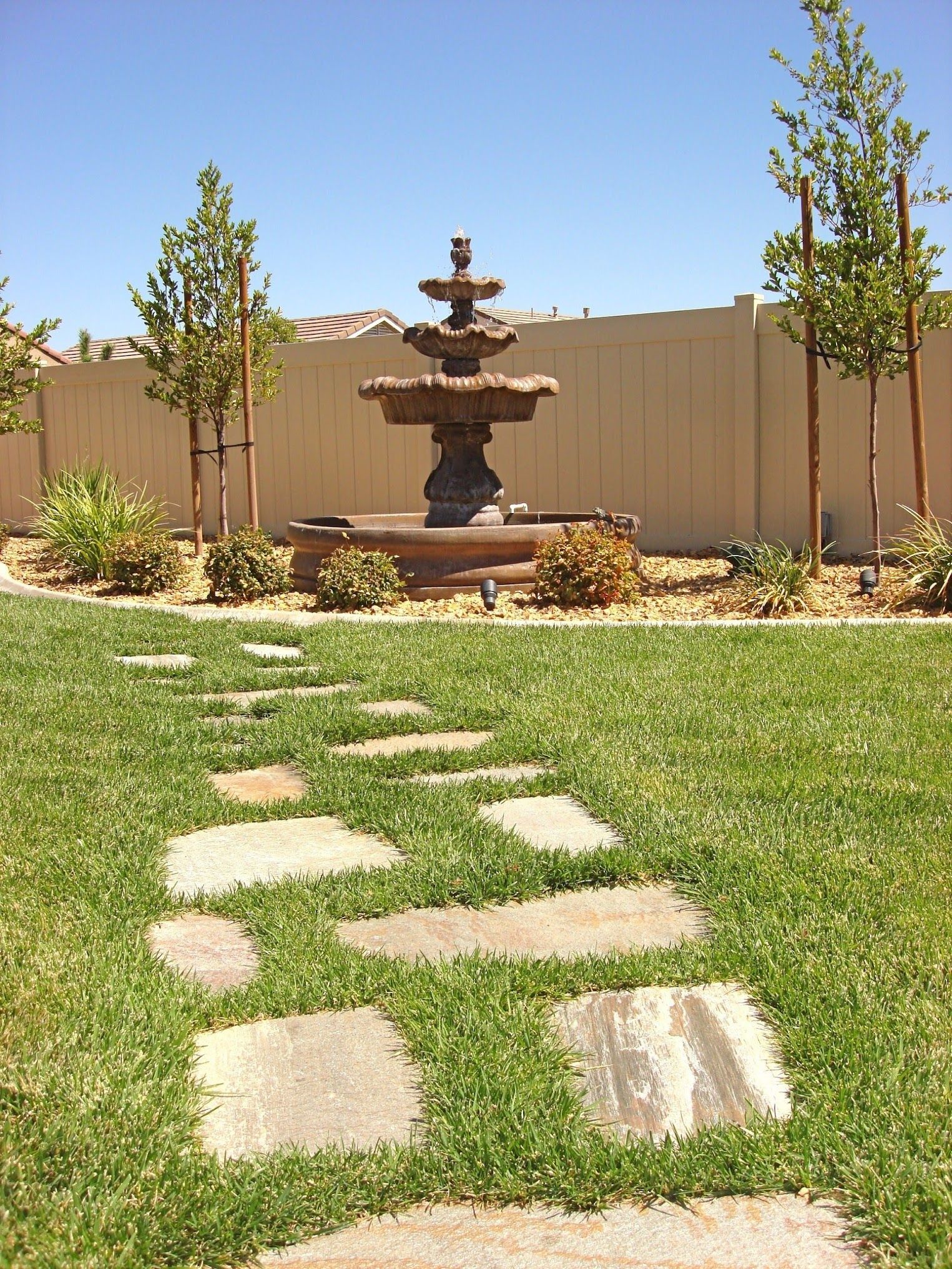 Stone path leads to a tiered fountain in a grassy yard, with trees and a beige fence in the background.