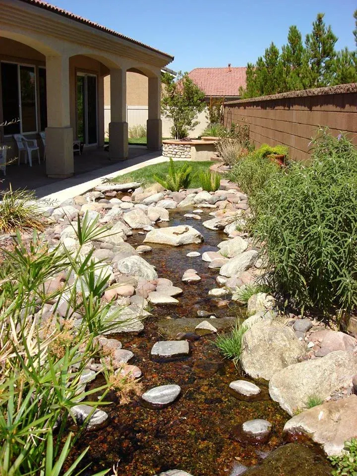 A decorative stream flows through a rock garden, beside a building with a patio, and a brown wall.