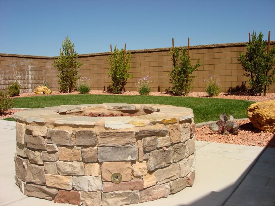 Stone fire pit in a backyard with green grass, small trees, and a beige block wall.