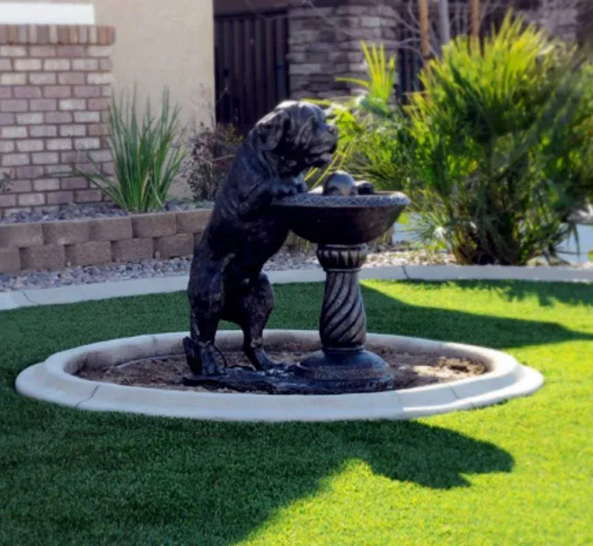 A dog statue drinking from a fountain in a green yard.