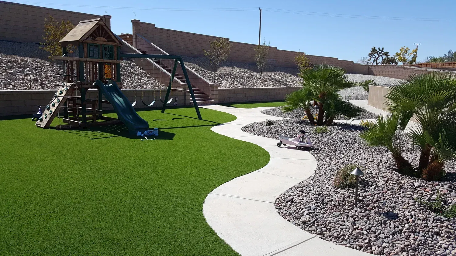 Playground with artificial turf, winding concrete path, rock landscaping, and a retaining wall under a blue sky.