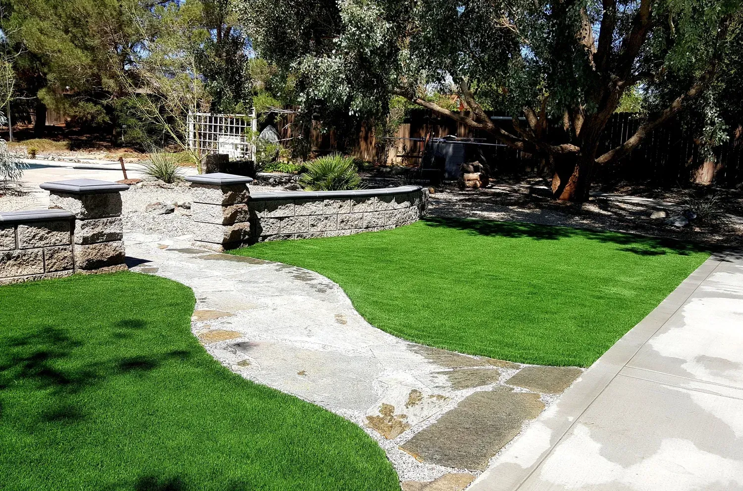 Green artificial turf and stone pathway in a backyard with trees and stone wall.