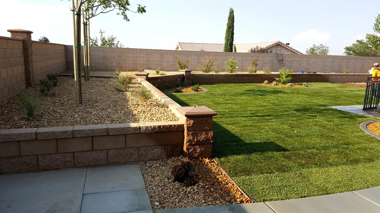 Backyard with retaining walls, gravel, grass, trees, and a concrete patio.