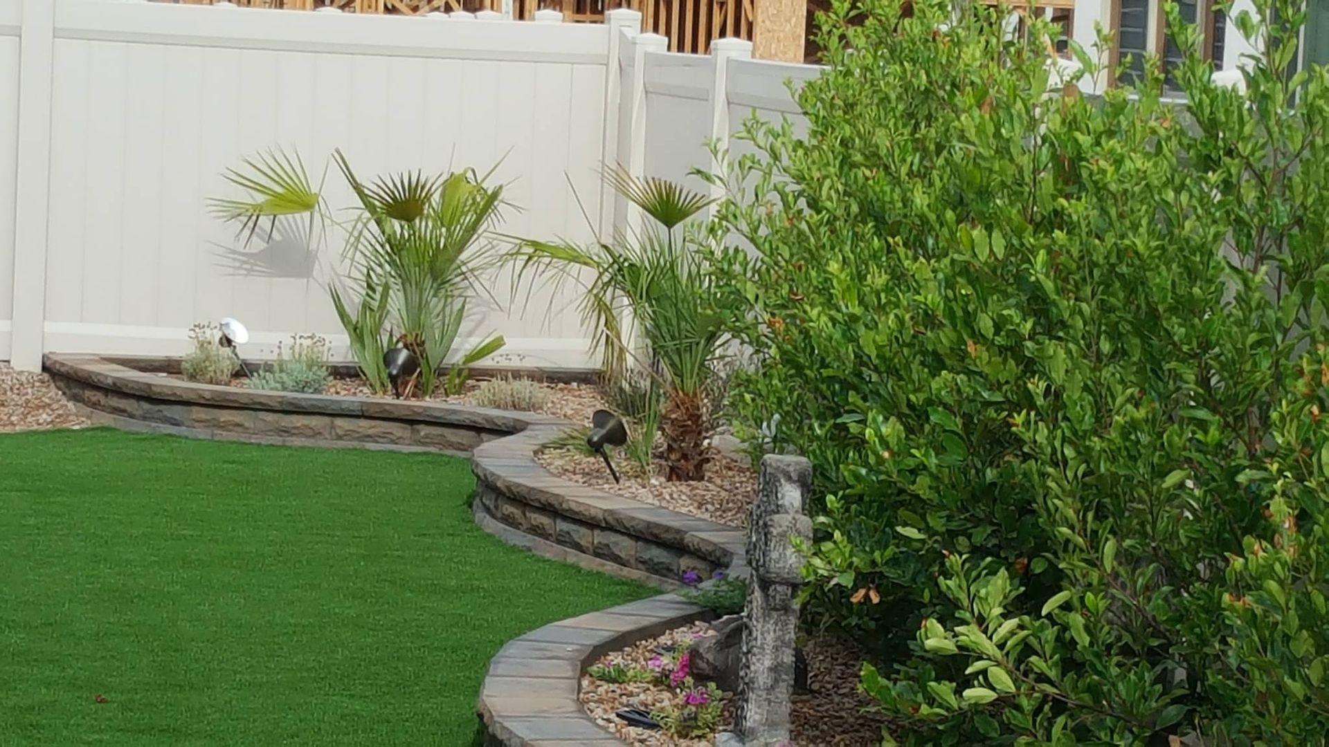 Backyard with artificial turf, curved retaining wall, and plants against a white fence.