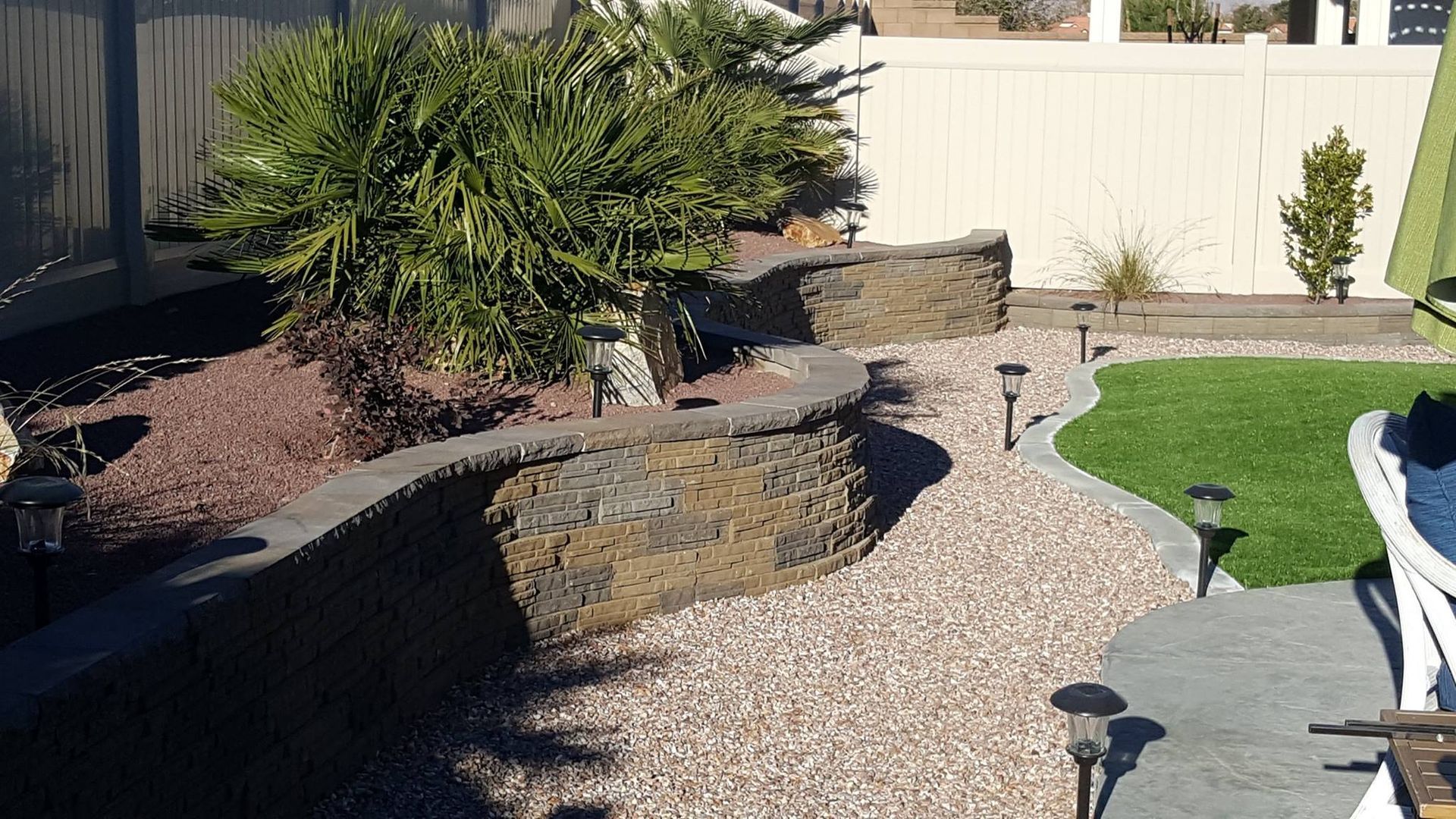 Backyard landscaping with rock walls, gravel, and artificial turf. Solar lights line the pathway.