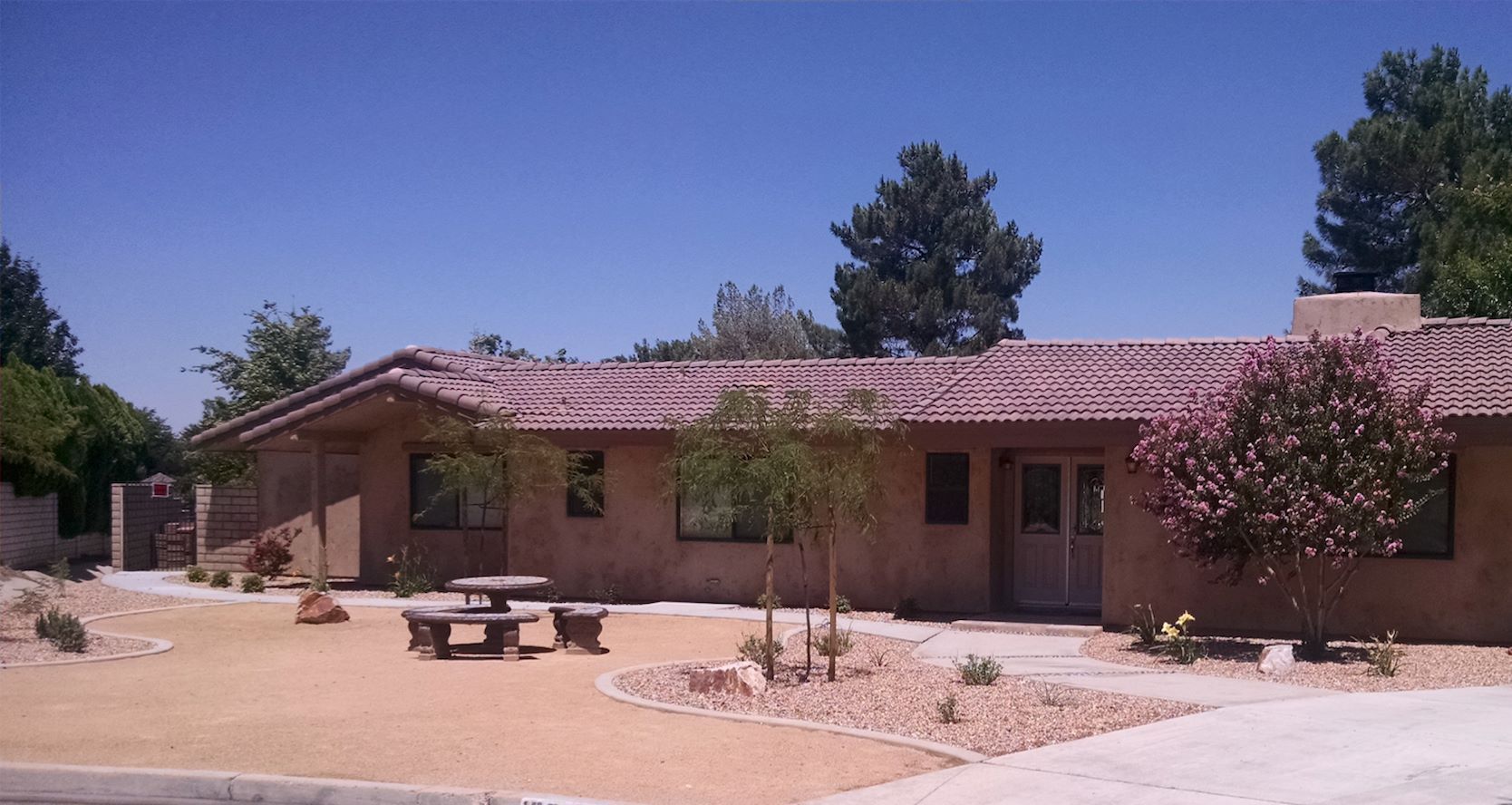 Tan house with red-tile roof, picnic table in front yard, surrounded by trees and shrubs, under a blue sky.