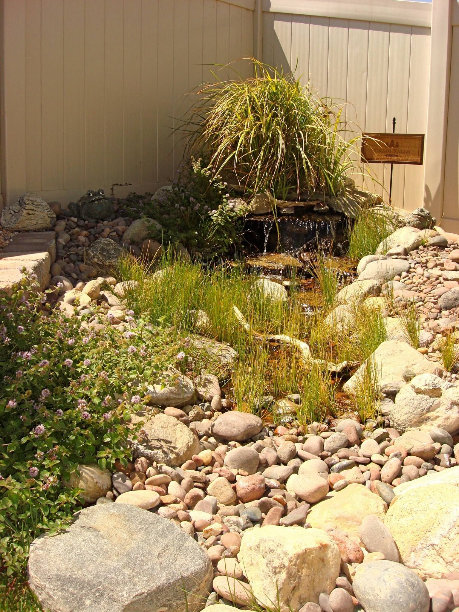 A small backyard water feature with cascading water and rocks, surrounded by greenery and a fence.