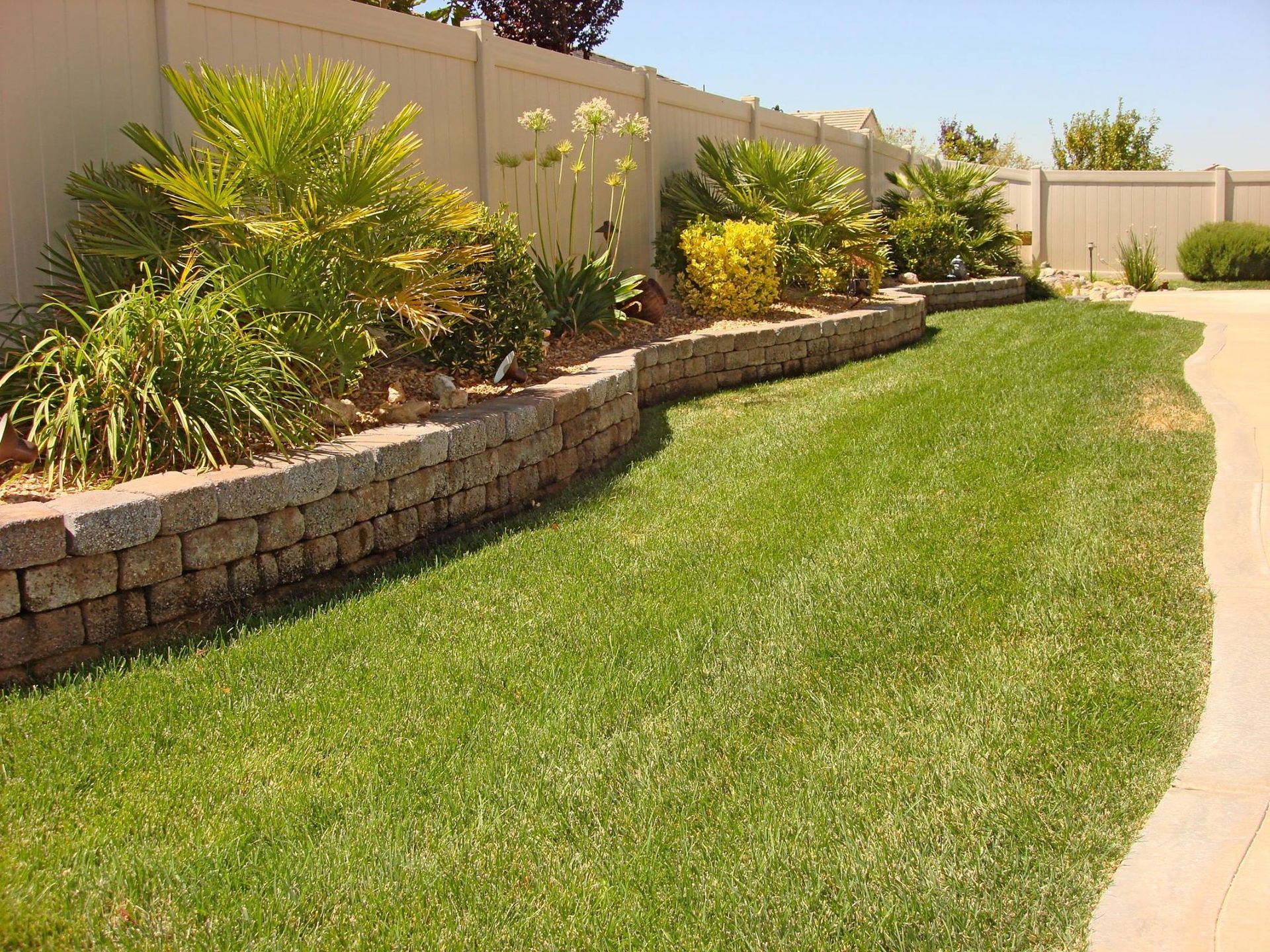 Green lawn with a retaining wall and plants, against a white fence.