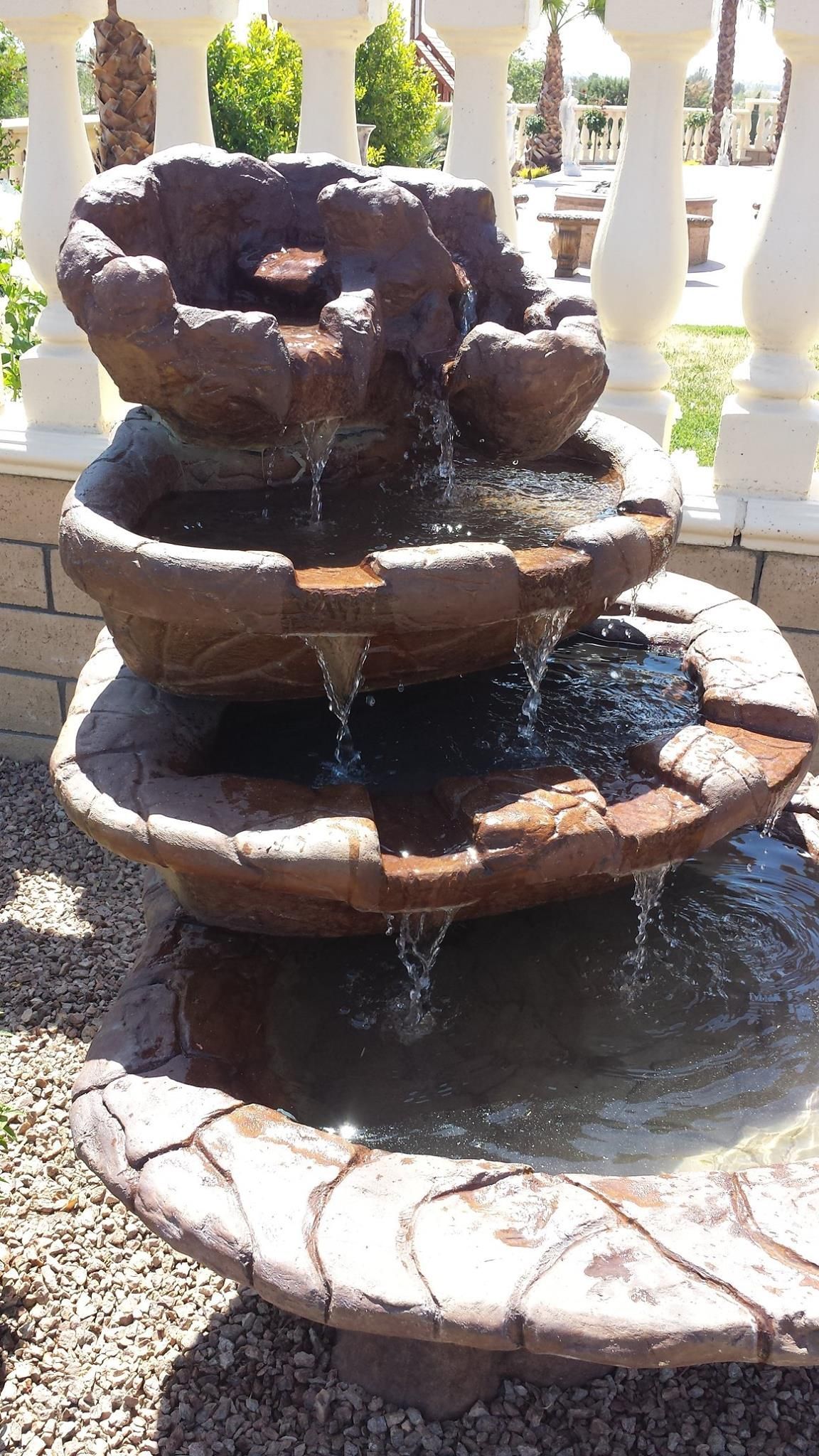 A tiered water fountain with water cascading down, set outside with a white railing in the background.