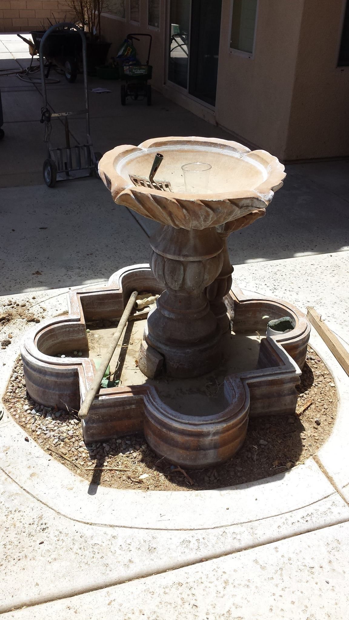 A stone fountain with a tiered base and upper bowl, set on a gravel bed in a yard.