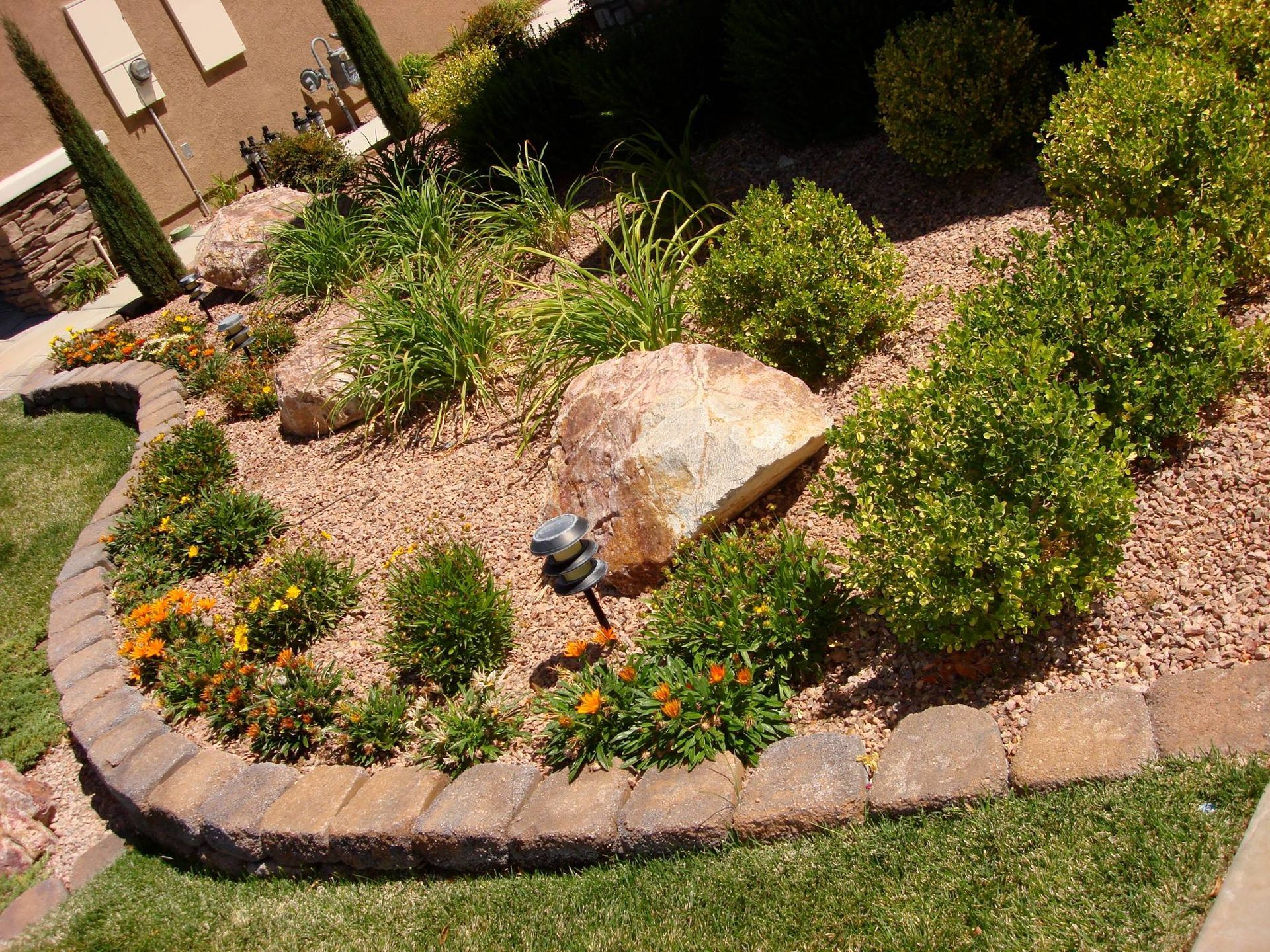 Landscaped garden bed with rounded brick edging, rock, small bushes, and orange flowers.