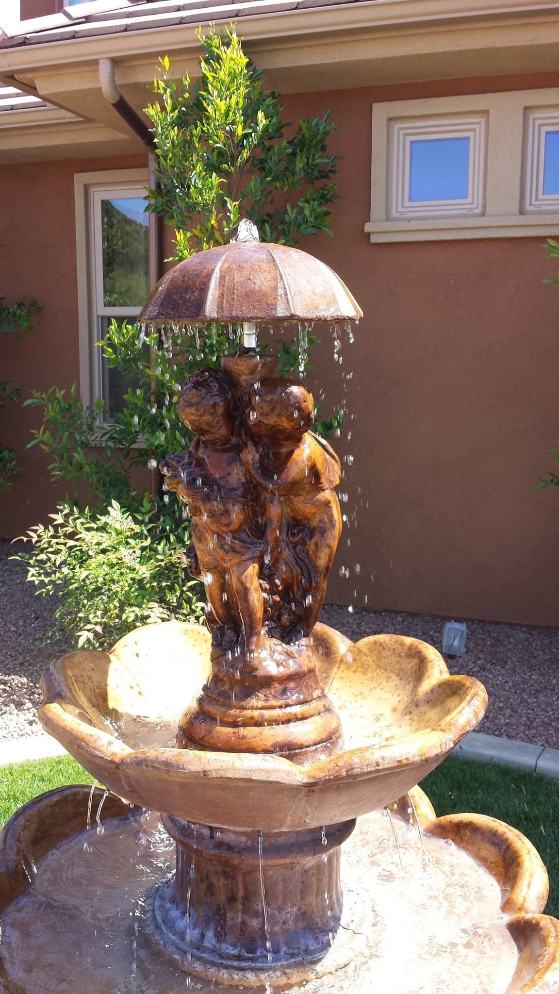 Water fountain with umbrella and cherub figures in a garden setting.