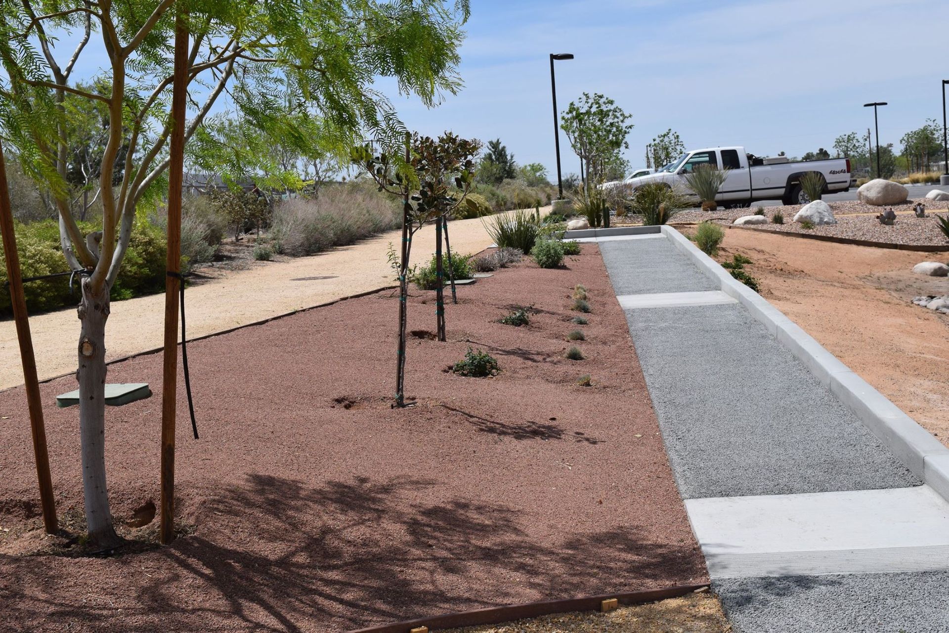 Landscaped area with gravel path, plants, and small trees next to a concrete walkway. A truck is parked in the background.