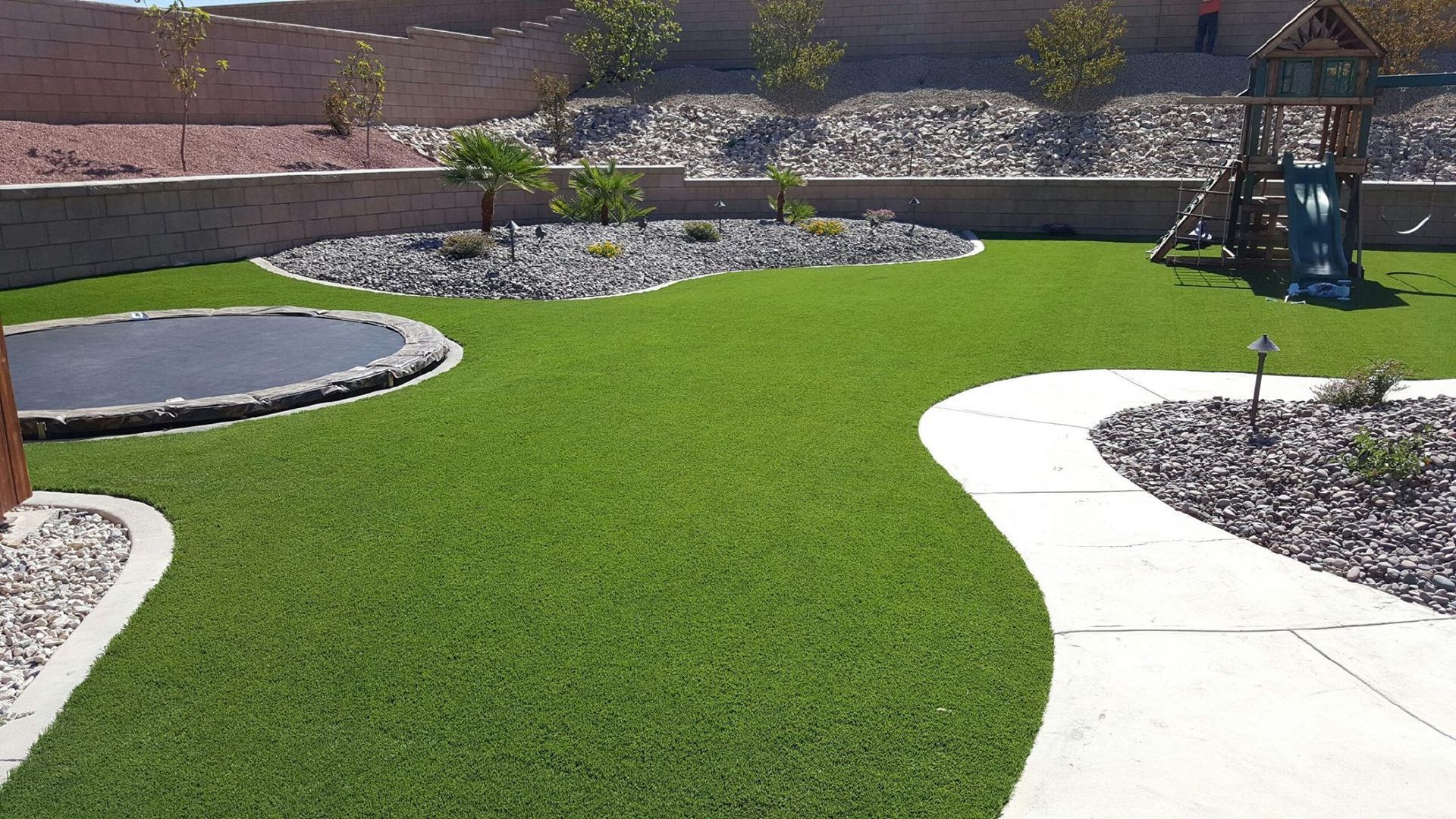 Green turf backyard with playground, trampoline, and rock garden beds.