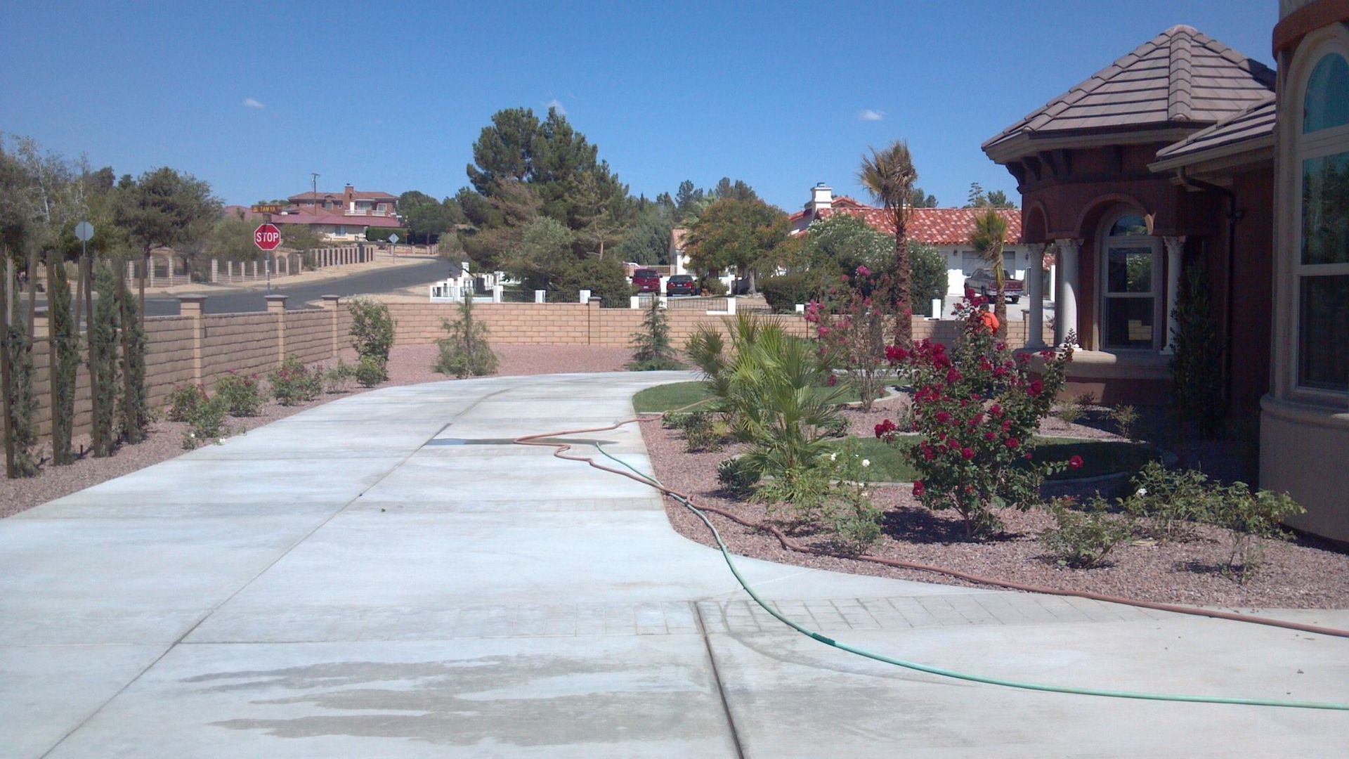 Driveway of a house with landscaping, fence, and neighborhood houses under a blue sky.