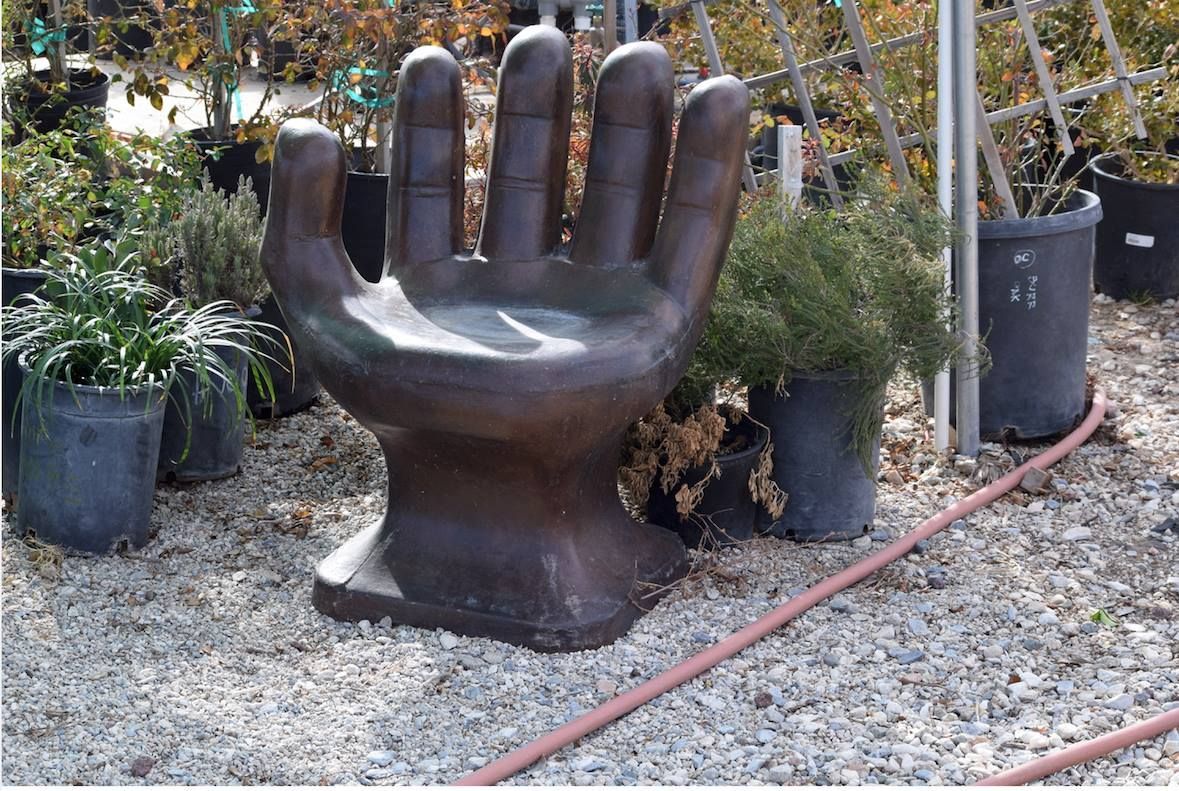 Bronze hand-shaped chair in a garden setting, surrounded by potted plants and gravel.
