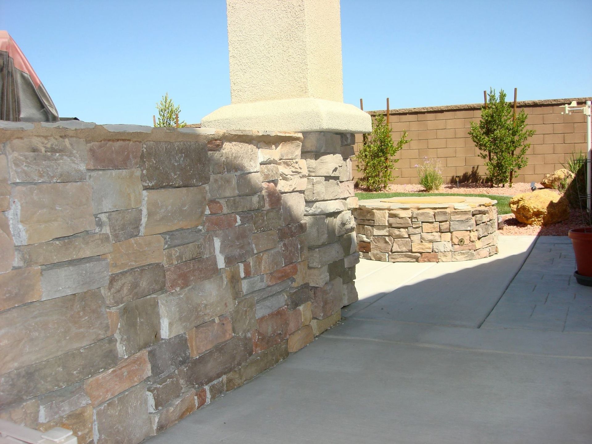 Stone wall and pillar on a patio, with a stone fire pit and a brick wall in the background. Sunny day.