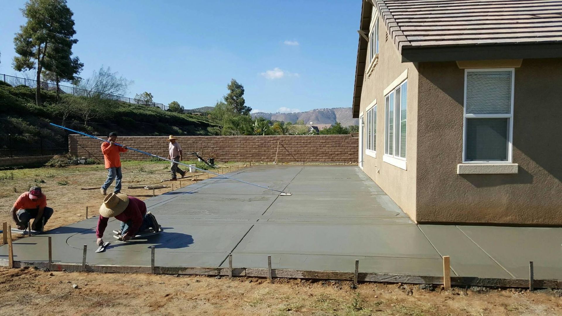 Construction workers pouring and smoothing concrete patio next to a house on a sunny day.