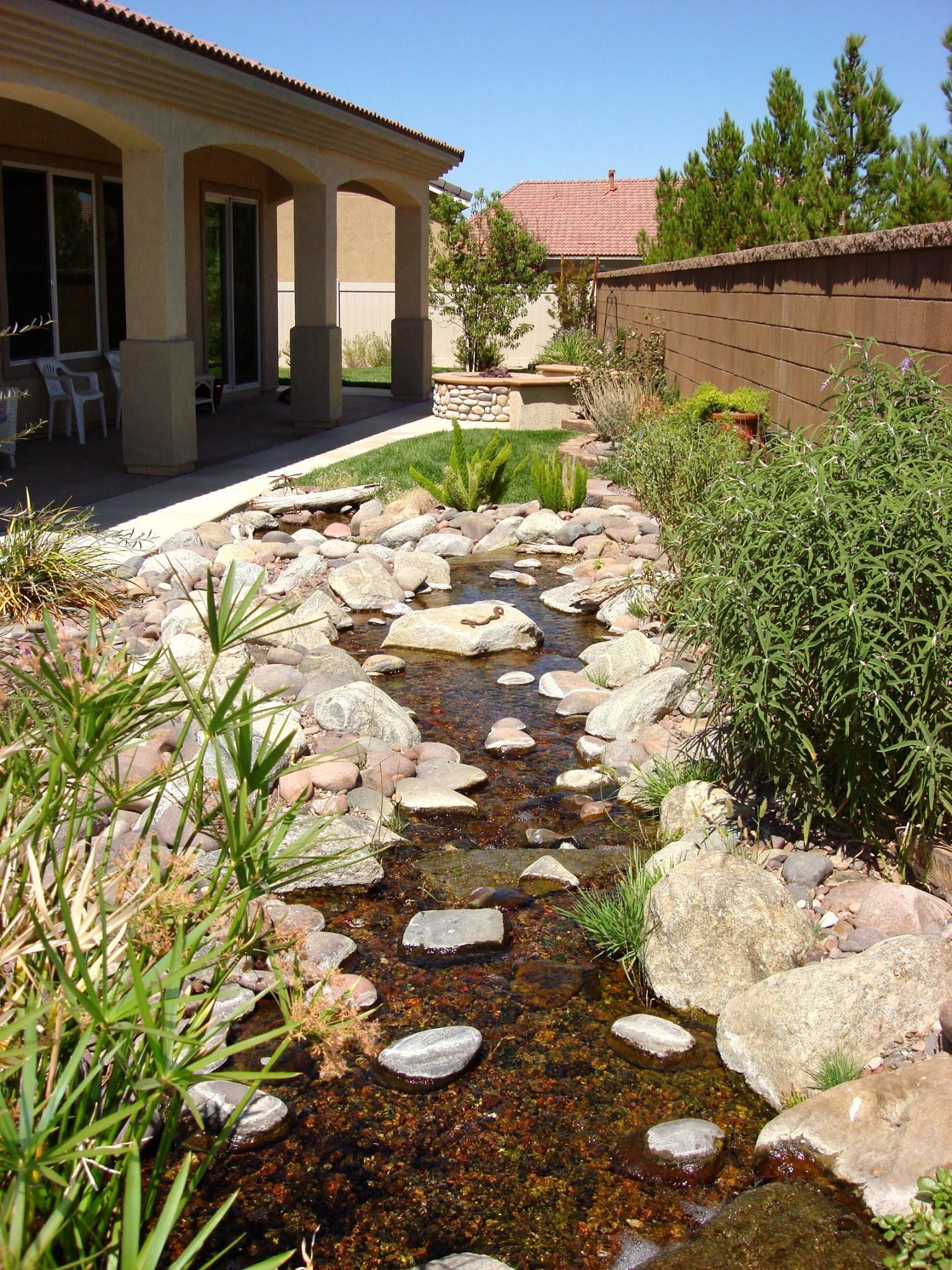 A shallow stream flows through a garden, alongside a covered patio and a brown wall.