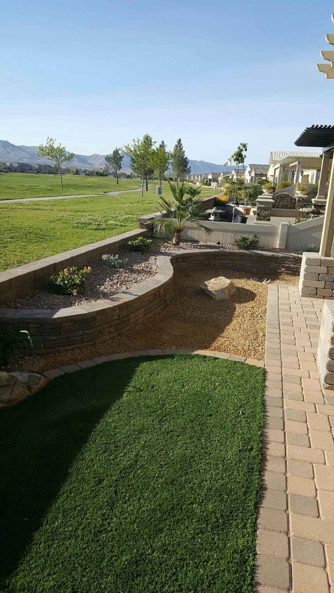 Lush green lawn alongside a rock garden, pathway, & low retaining walls with a golf course in the background on a sunny day.