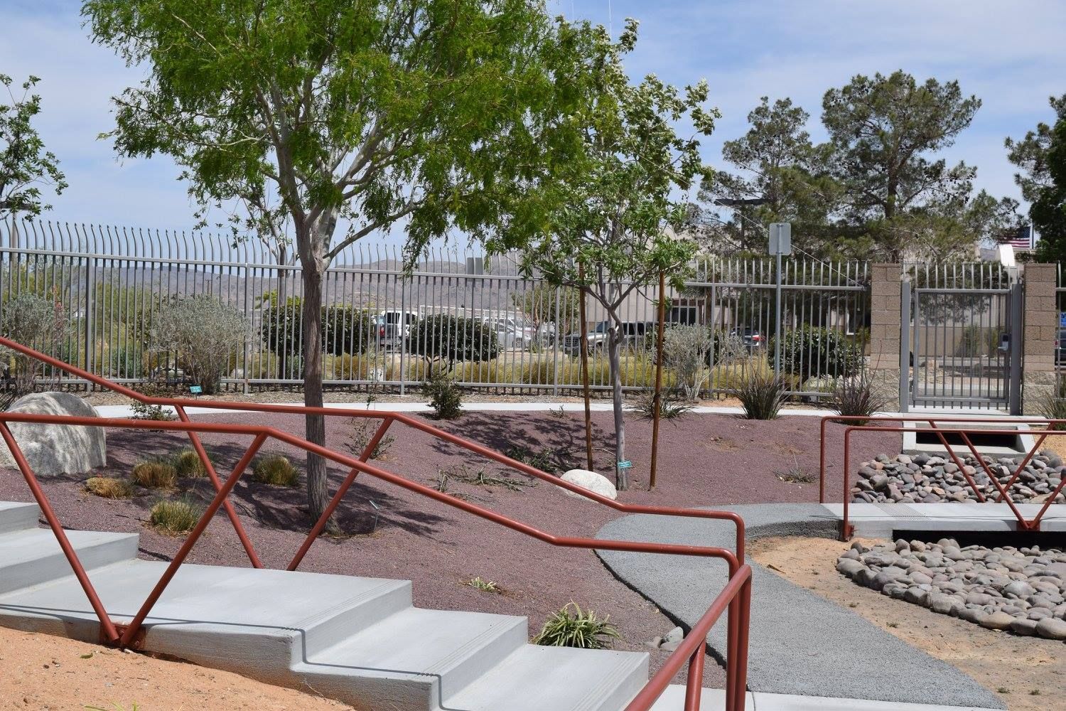 Concrete steps with red handrails leading to a landscaped area with trees, gravel, and a fence.