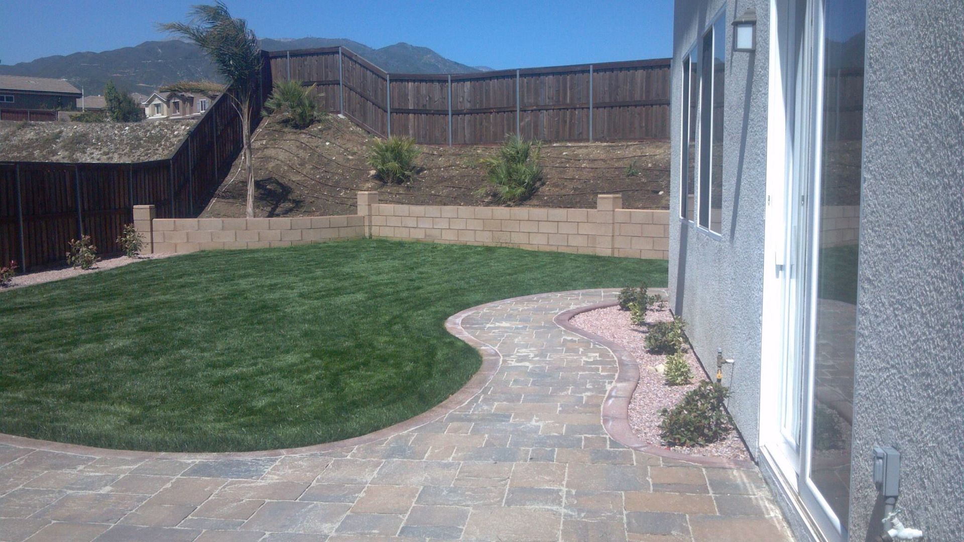 Backyard with lawn, stone path, retaining wall, wooden fence, house, and mountains.