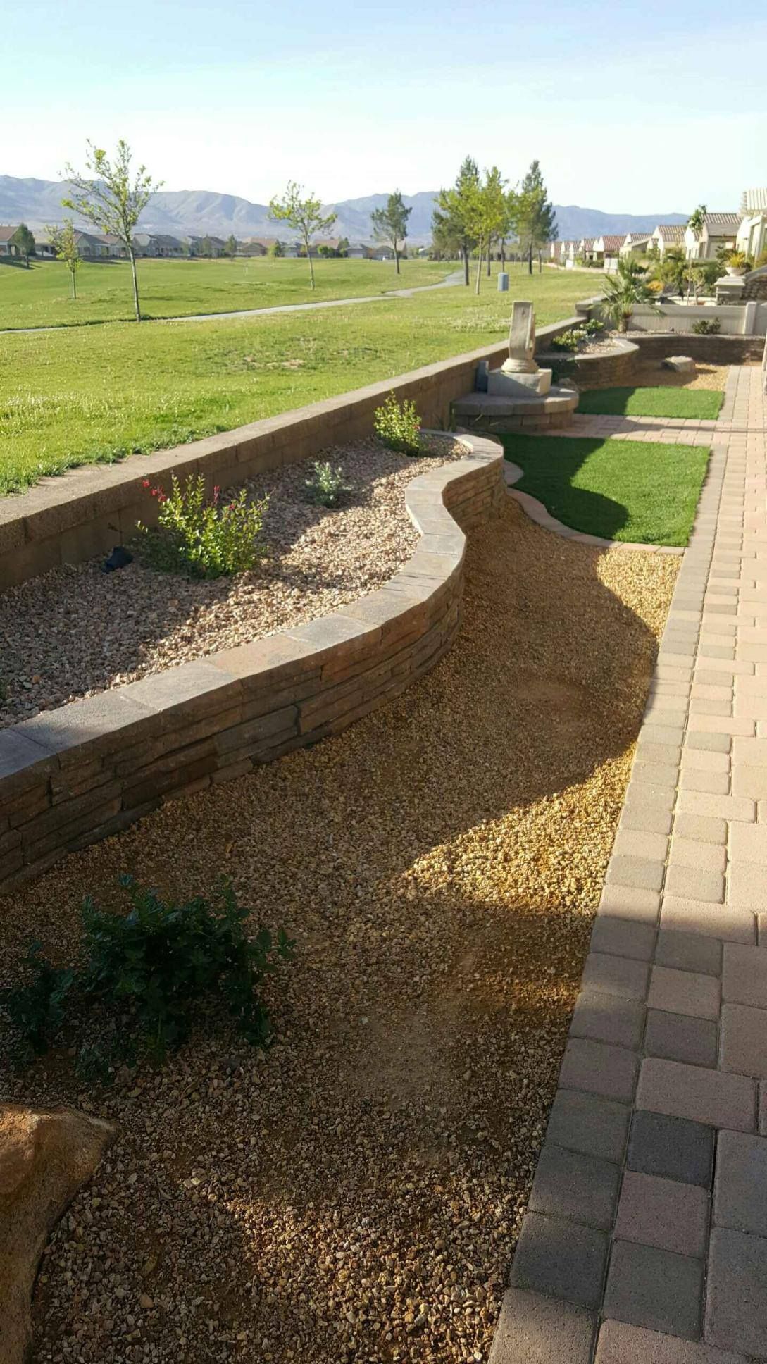 Stone-lined landscaping feature with gravel, plants, and retaining walls, leading to a grassy area and distant hills.