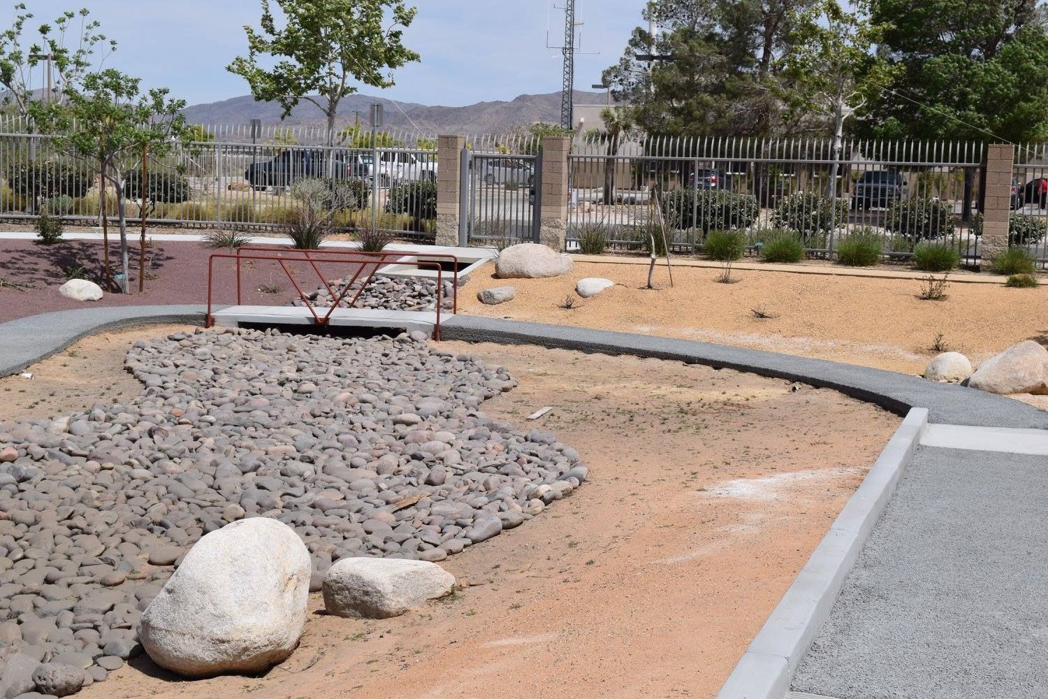 Park with gravel paths, rock features, and a small bridge. Fenced perimeter with sparse vegetation and distant buildings.
