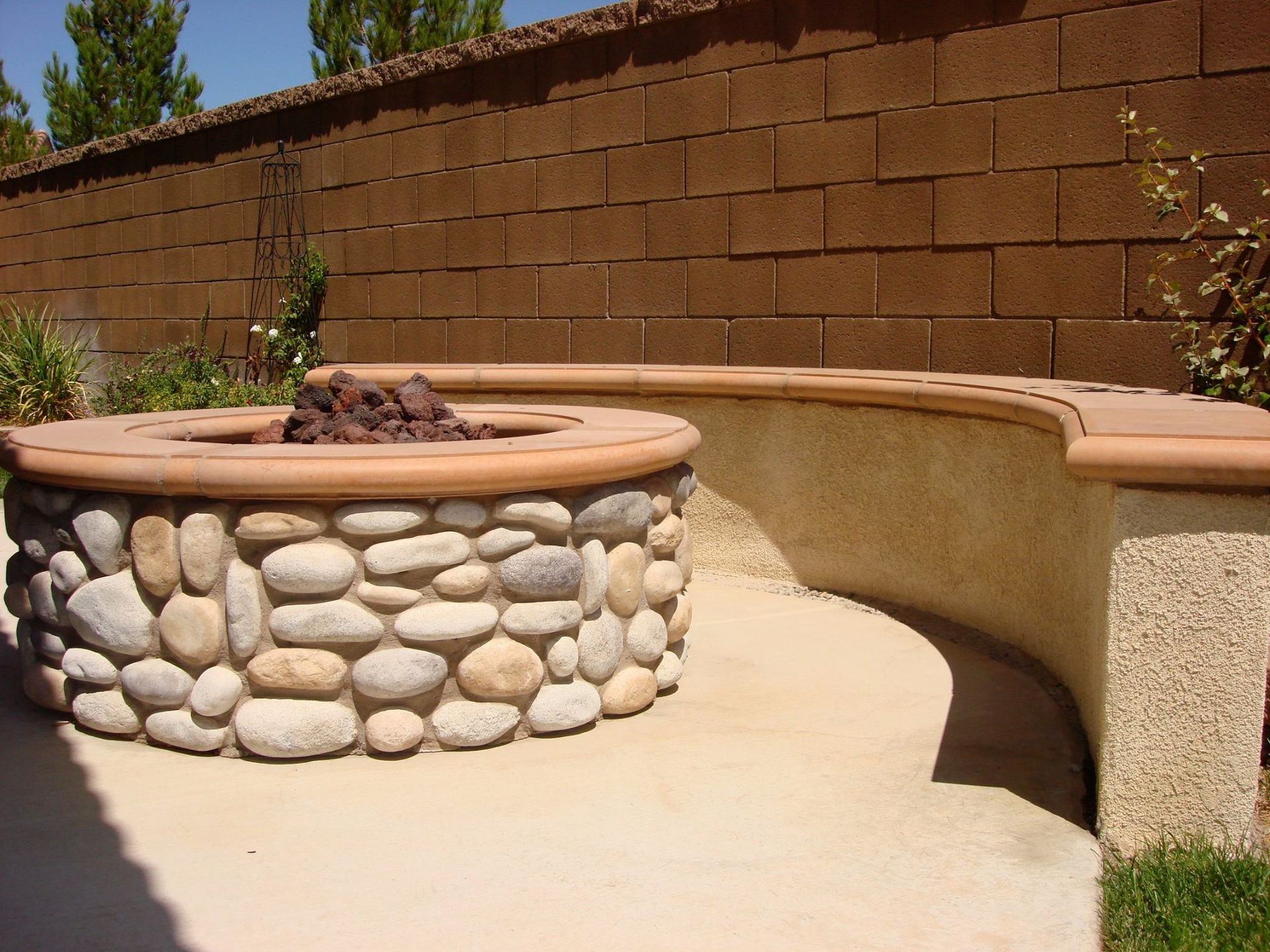 Fire pit with stone exterior and curved bench on a patio, set against a brown brick wall.