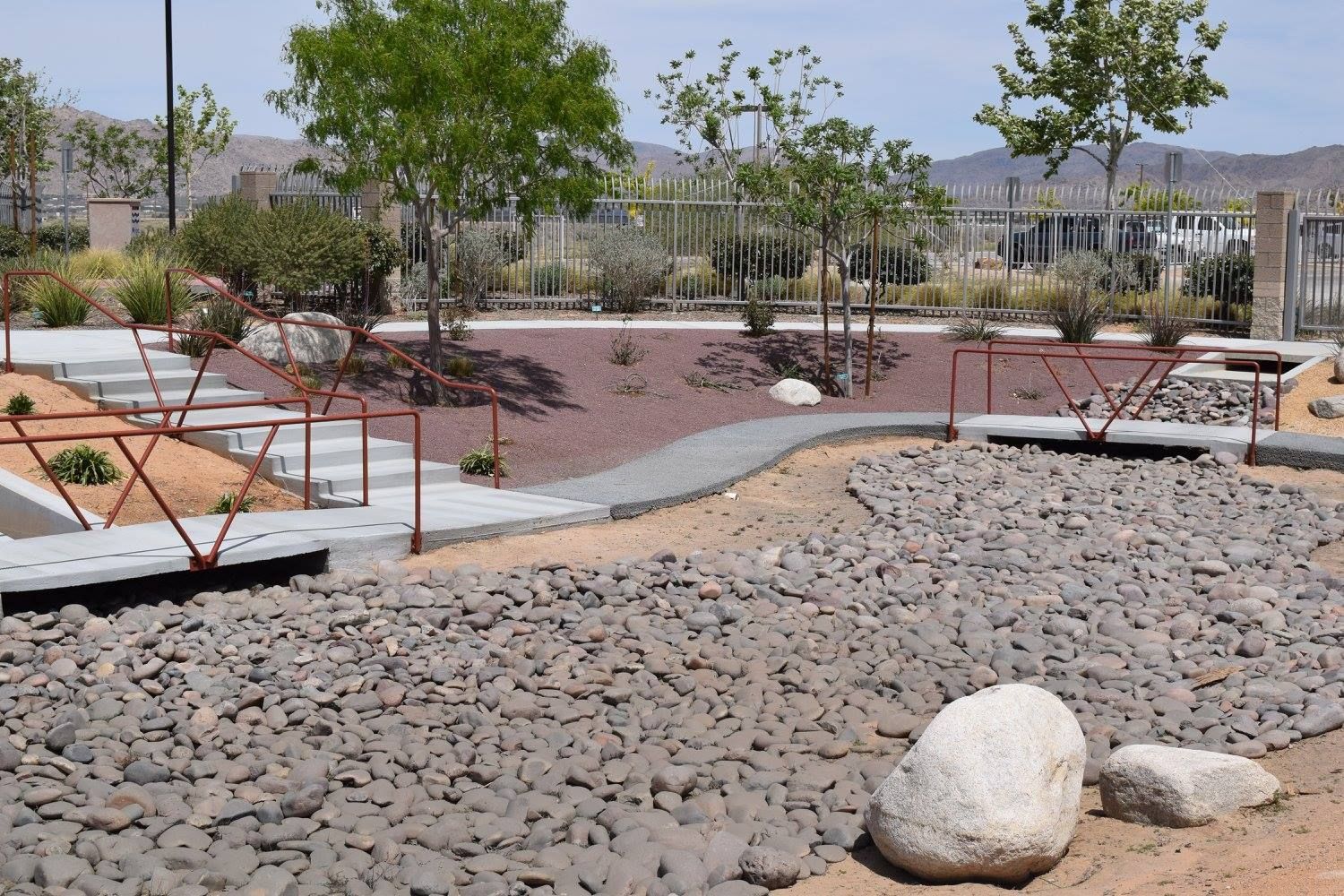 Dry creek bed with rock pathway, stone steps, and small red metal bridges in a park-like setting with trees.