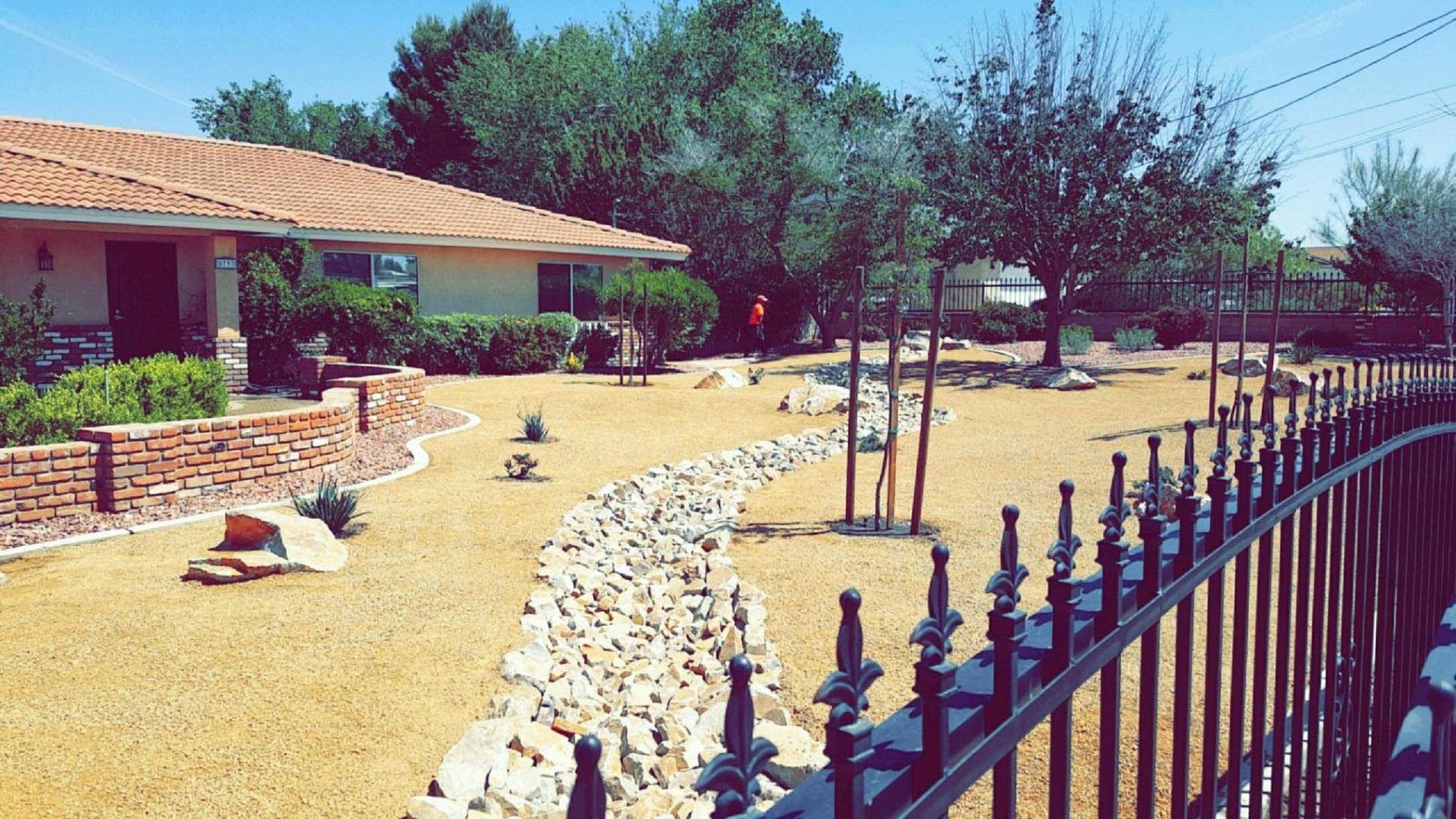 Tan house with terracotta roof, landscaped front yard with gravel, rocks, trees, and black fence.