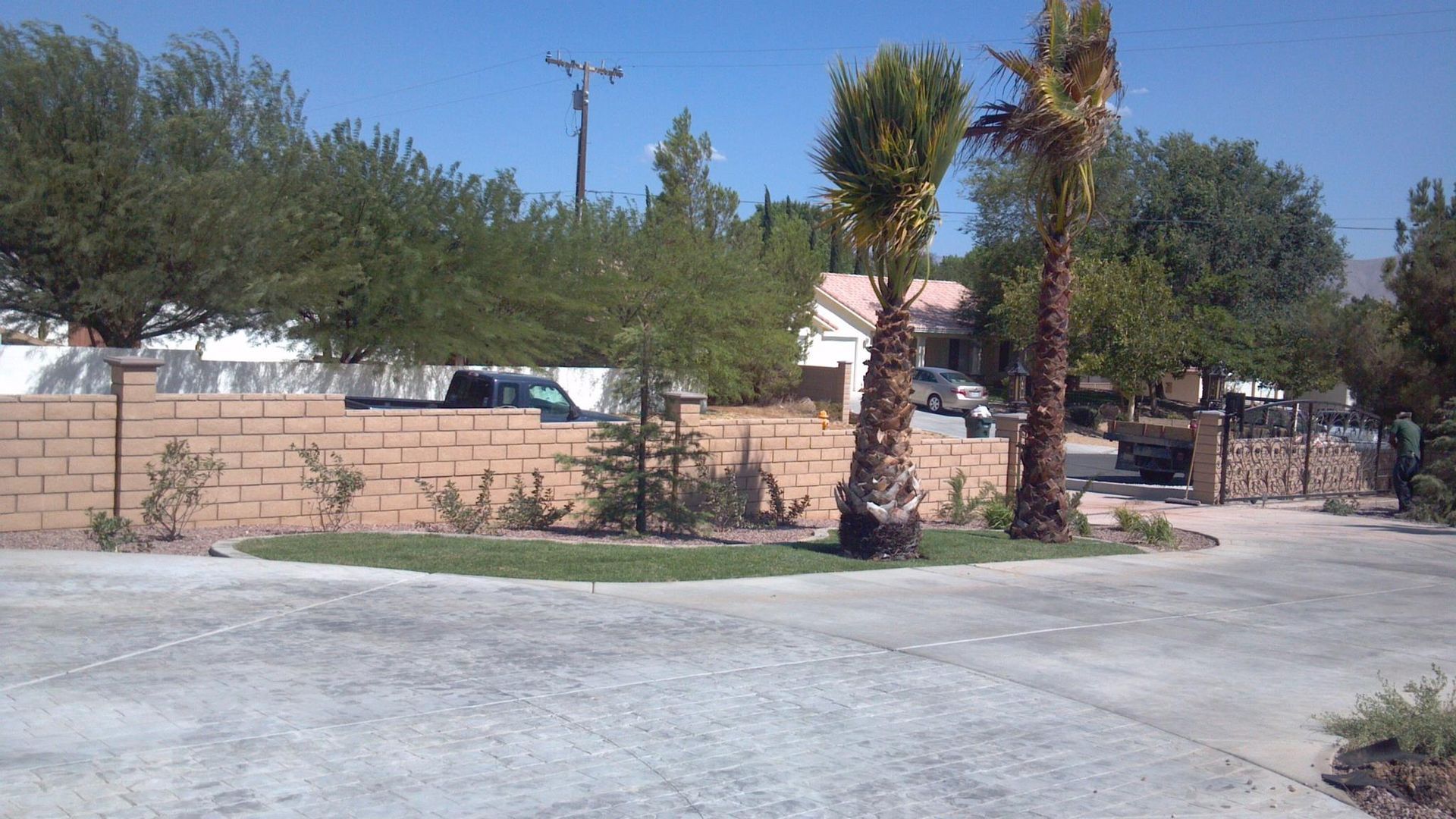 Brick wall and driveway leading to a house with trees and palm trees under a blue sky.