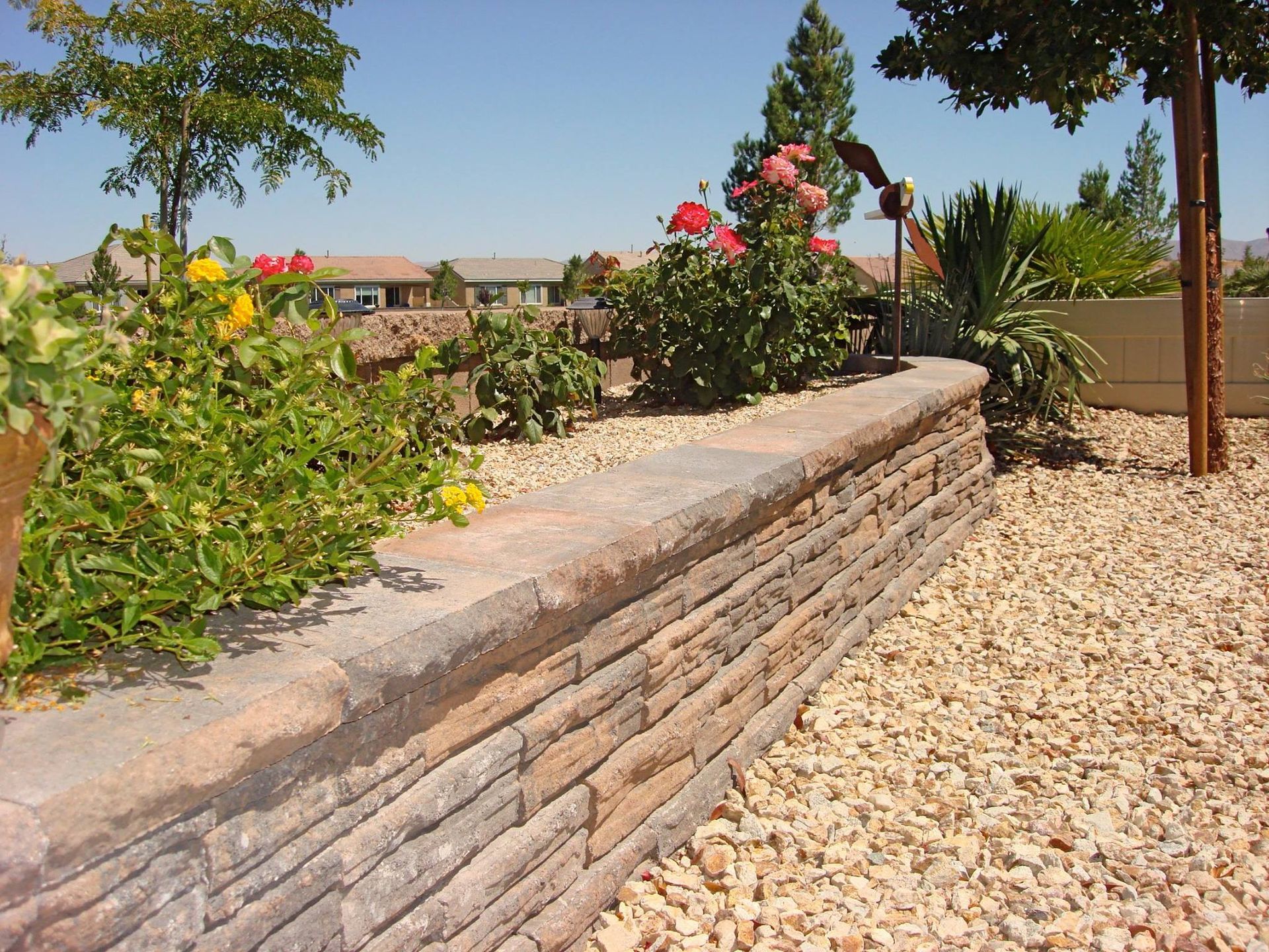 Low stone retaining wall with a flower bed on top, surrounded by gravel.