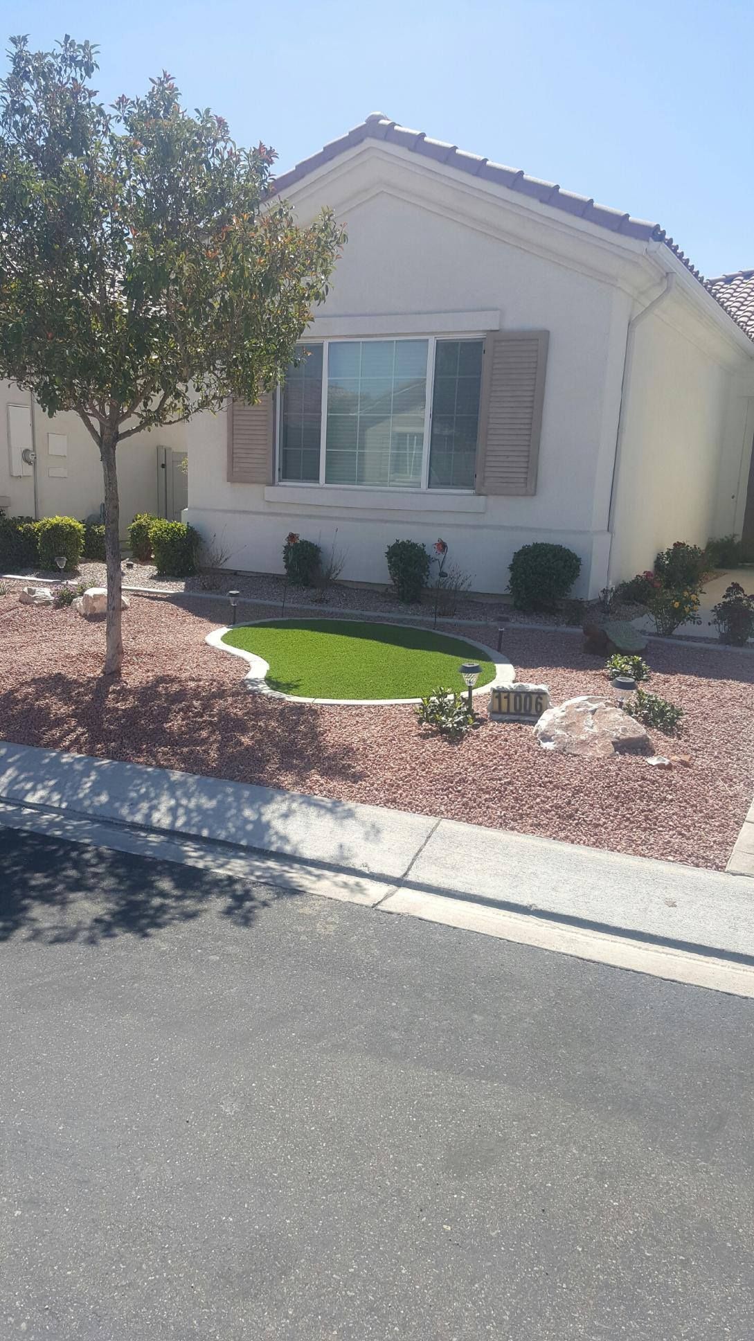 House exterior with tan siding, red rock landscaping, small lawn, tree, and blue sky.