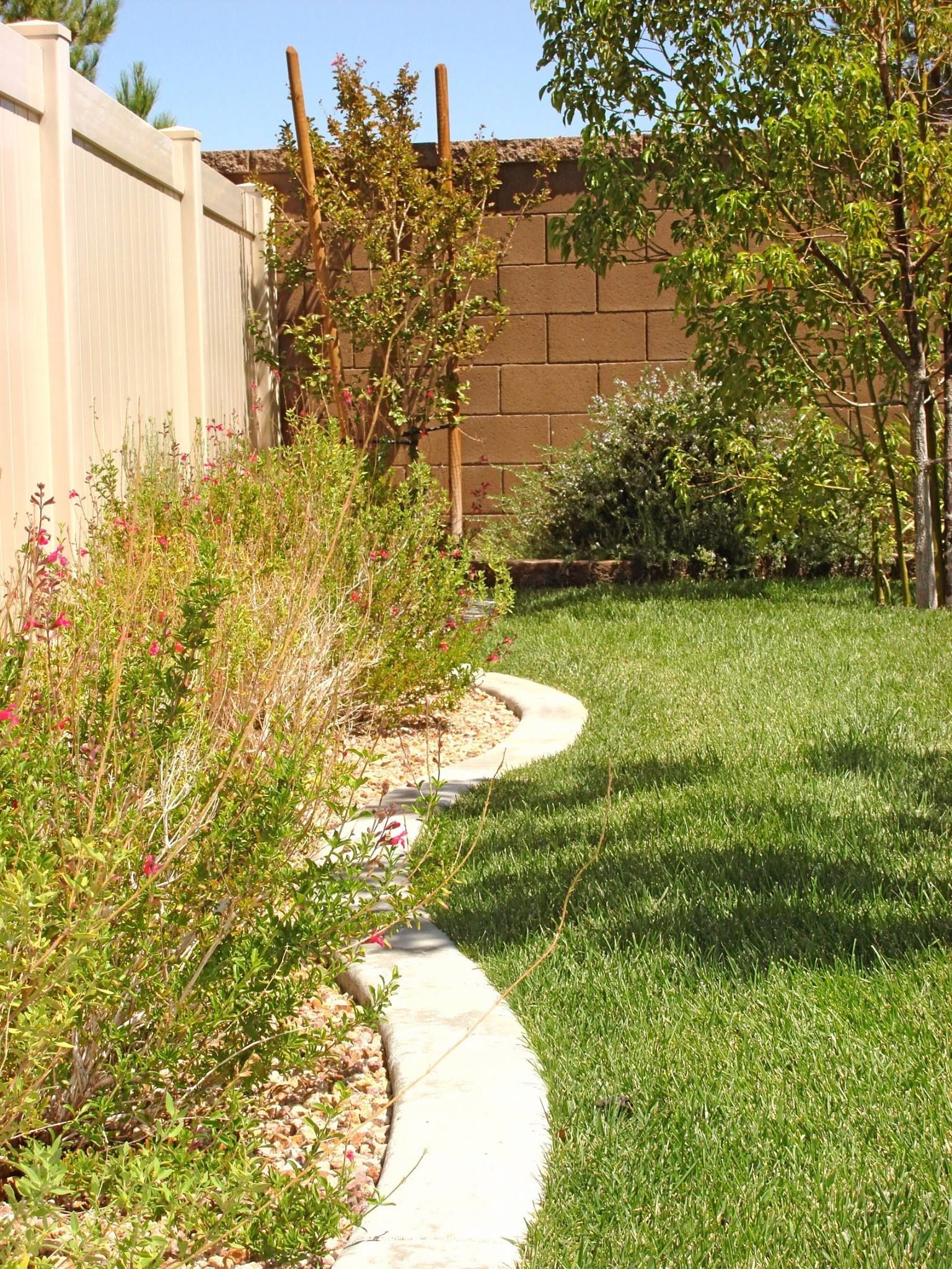 Backyard with a lawn, flower beds, concrete border, and a tan fence.