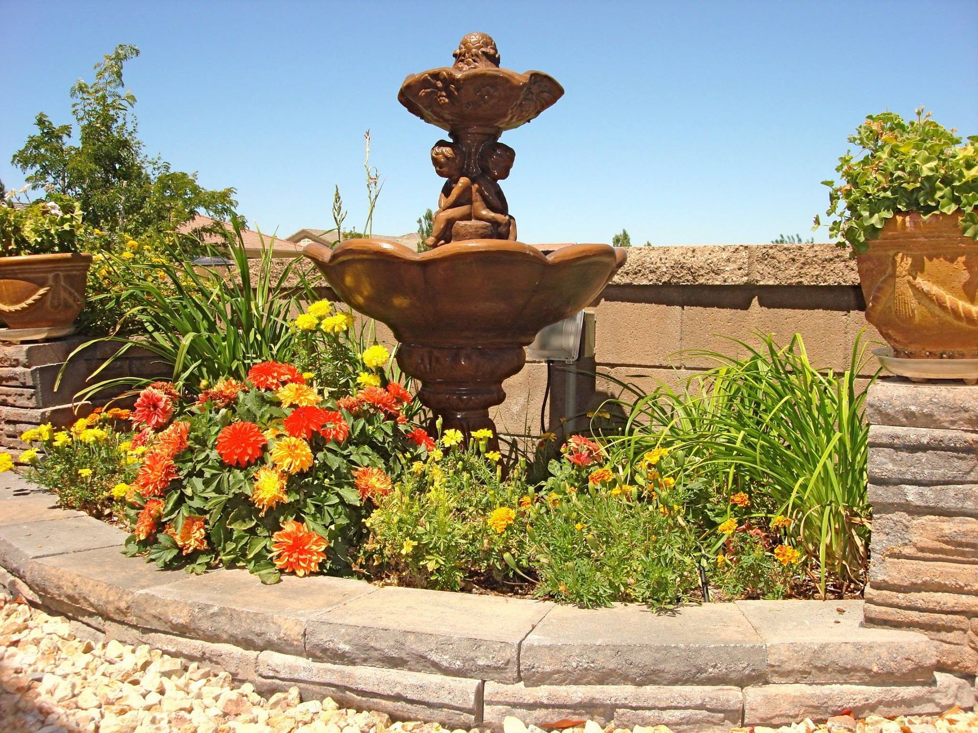 Stone fountain in a garden bed with orange and yellow flowers, blue sky background.