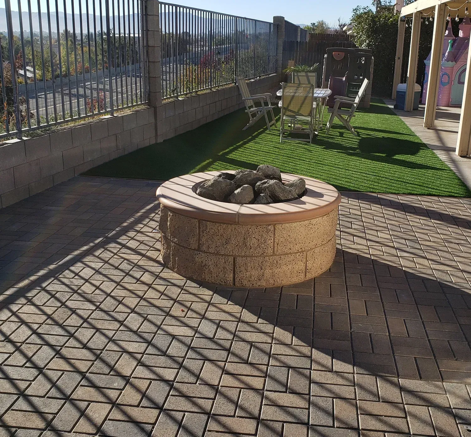 Brick fire pit on a patterned patio with artificial grass and seating in a backyard.