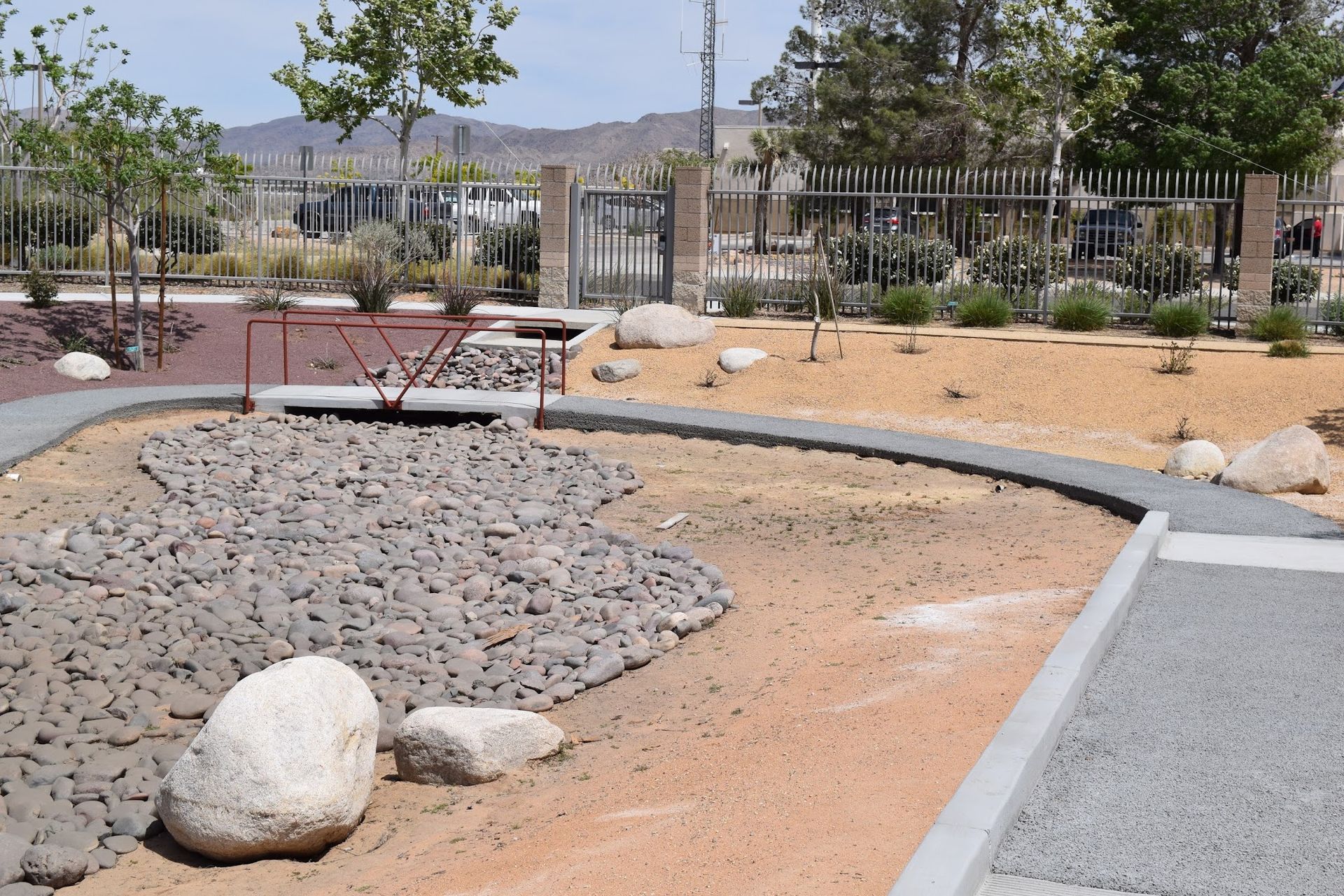 A dry landscape with a path, rocks, gravel, and a metal bridge near a fence.
