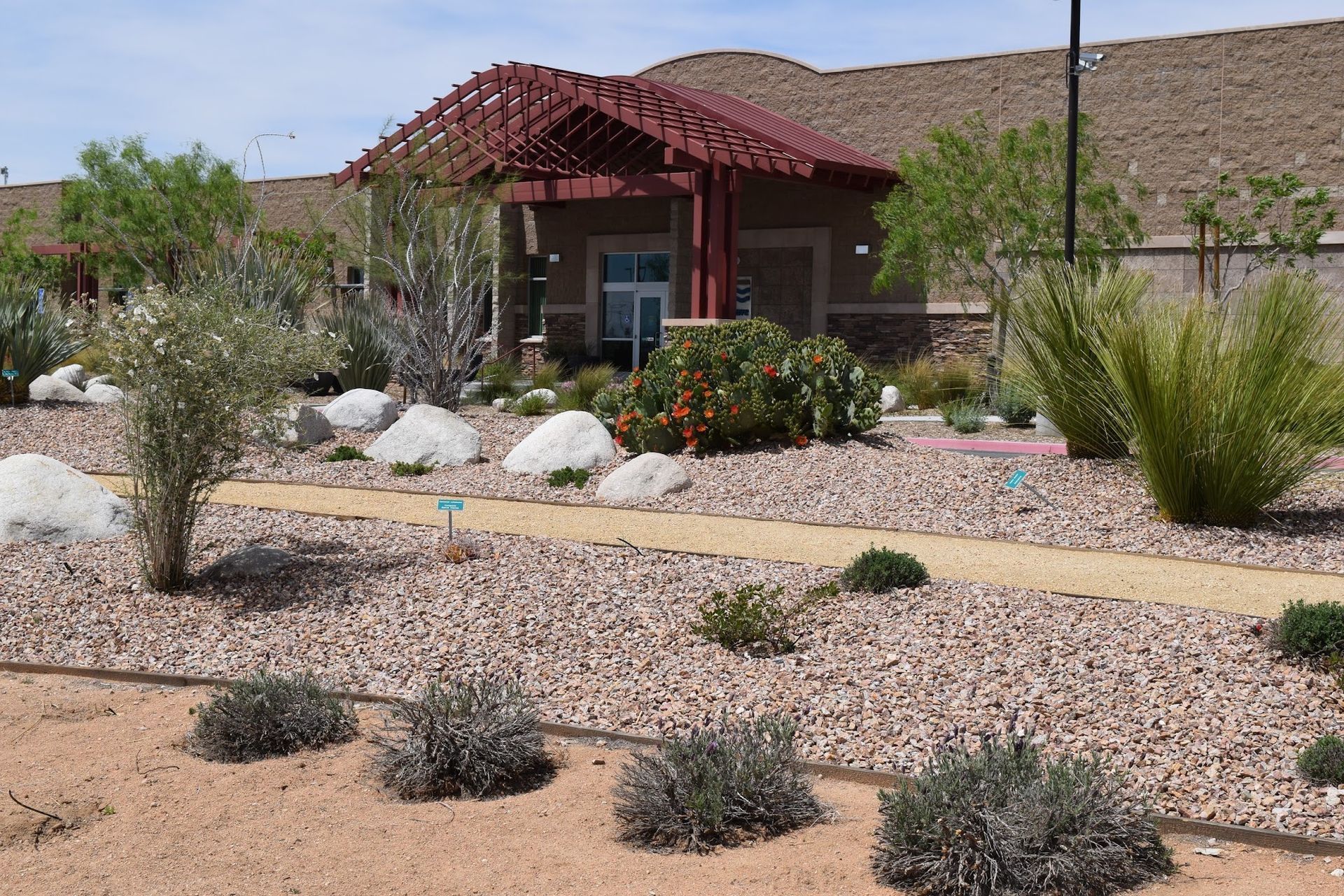 Desert landscaping with a small building, gravel path, rocks, and various plants. Red roof on building.