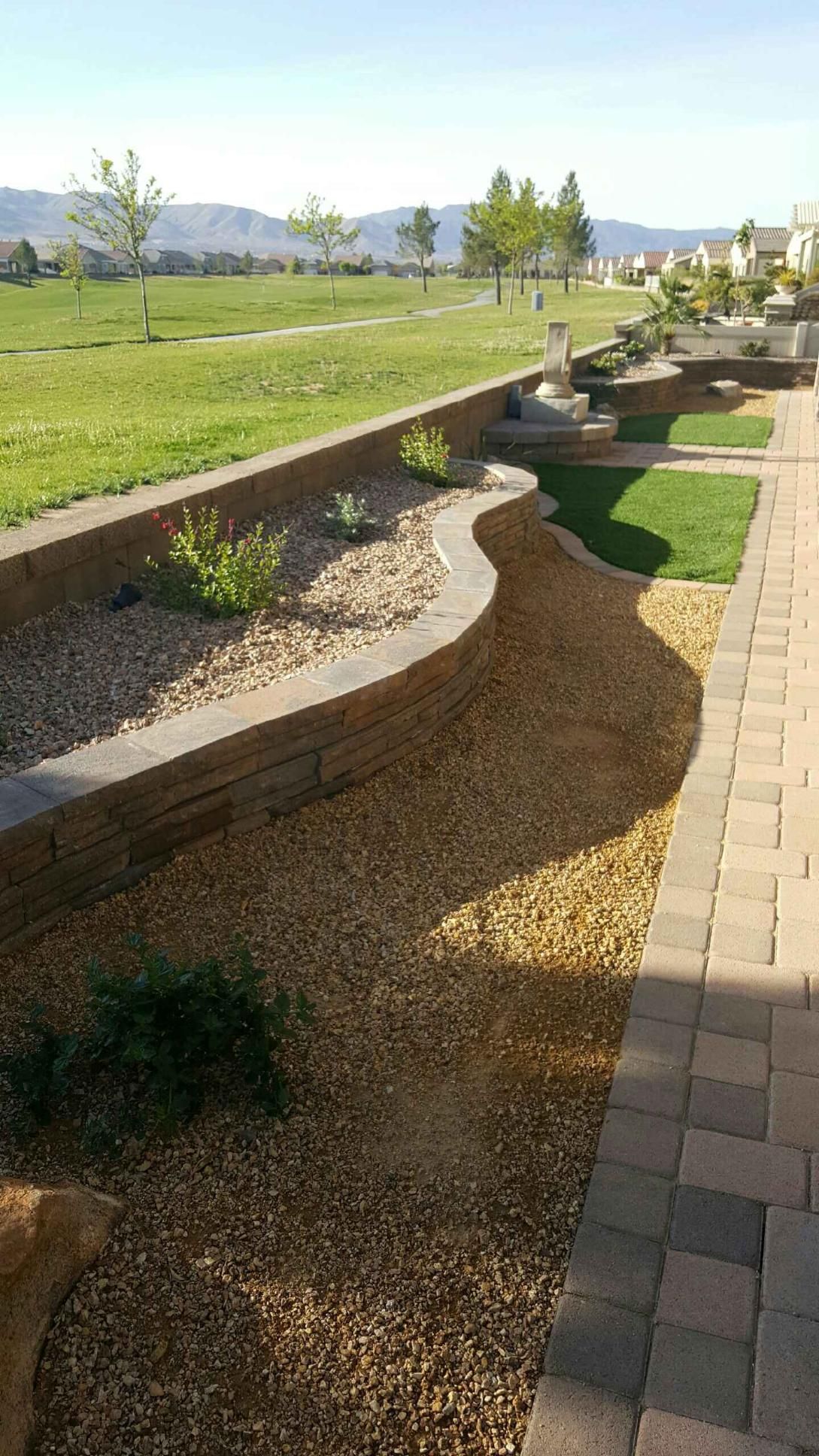 Curving landscaped area with gravel, small plants, and retaining walls, next to a brick patio and lawn.