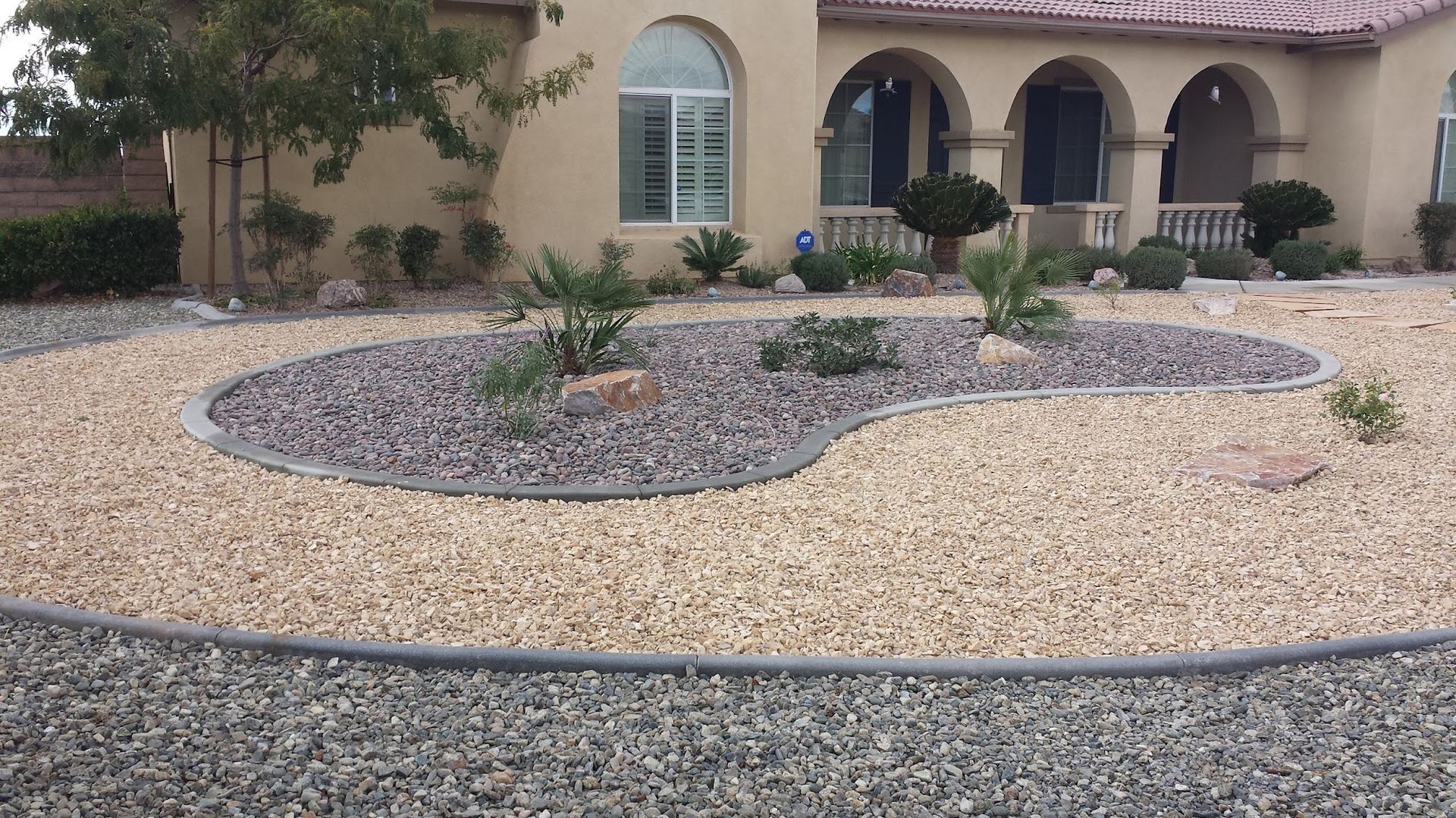 Front yard landscaping with rock beds and curved borders in front of a tan house.