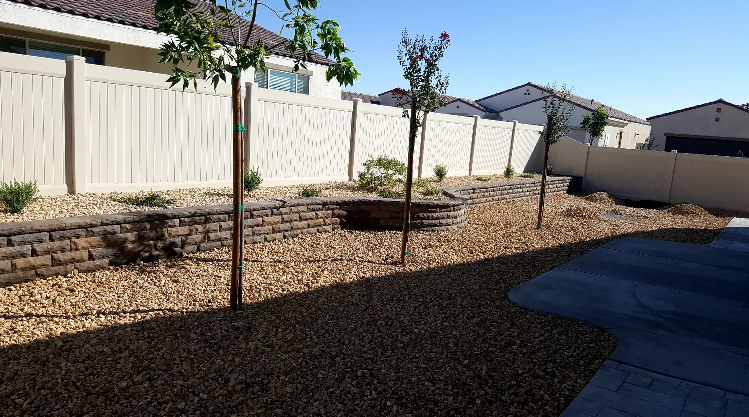 Backyard landscaping with retaining wall, trees, gravel, and beige fence. Sunny day.