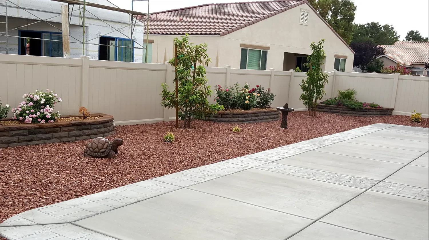 Landscaped yard with red rock, flowers, trees, and a beige fence in front of a house.