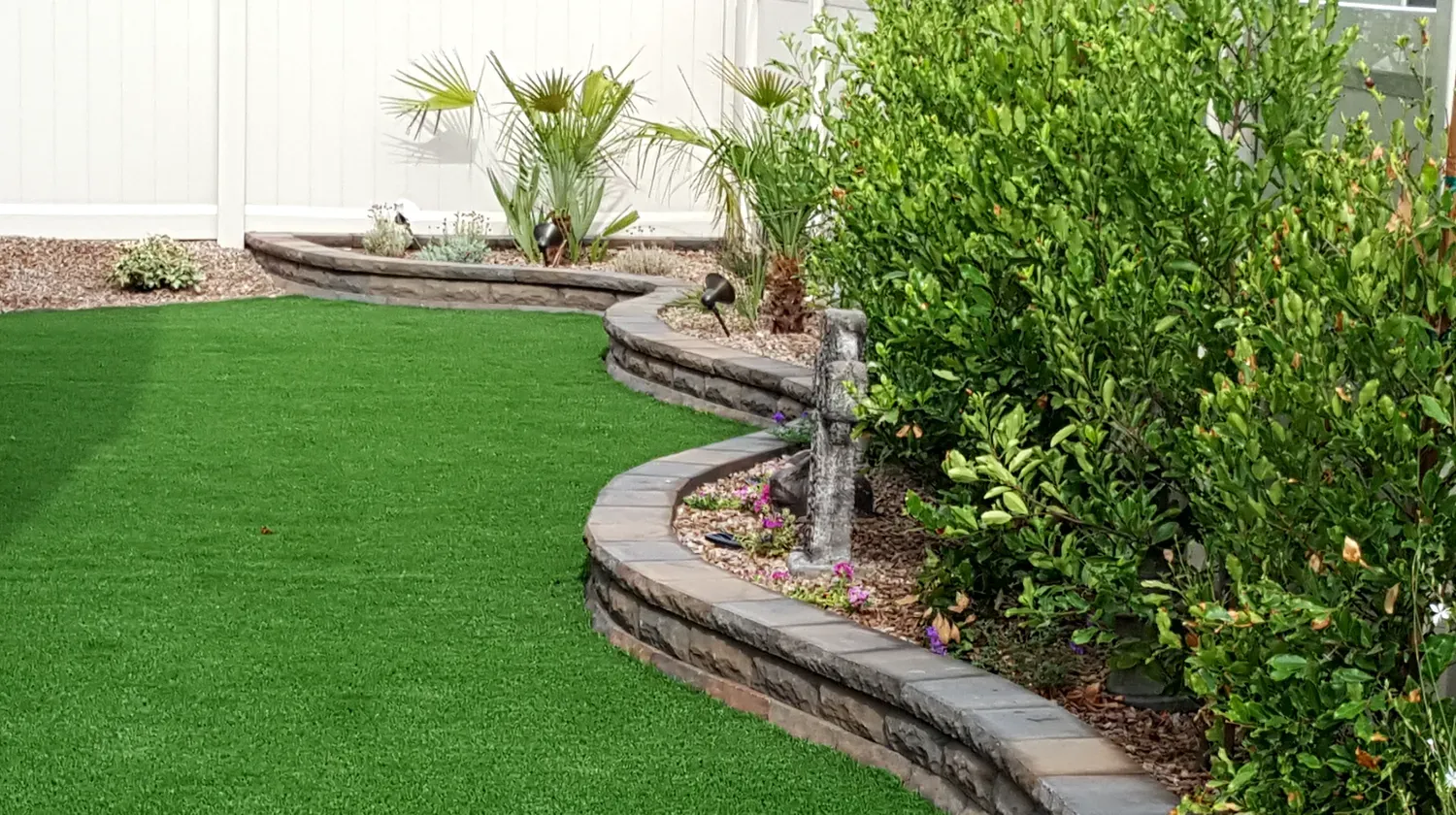 Green lawn bordered by a curving stone wall with plants and a white fence backdrop.
