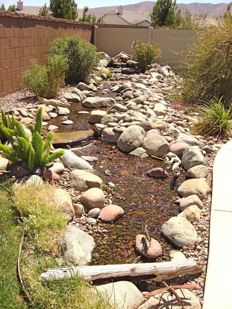 A backyard rock stream with a waterfall, surrounded by plants and a brick wall.