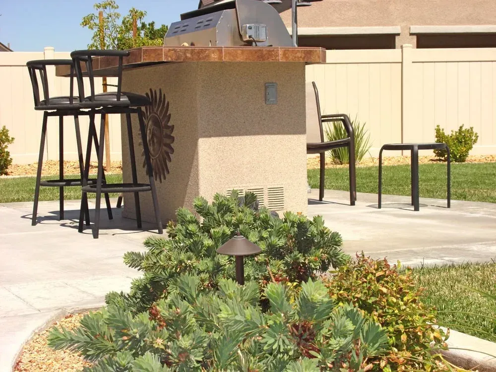 Outdoor kitchen with grill, seating, and landscaping; tan stucco structure, black chairs, and green plants.