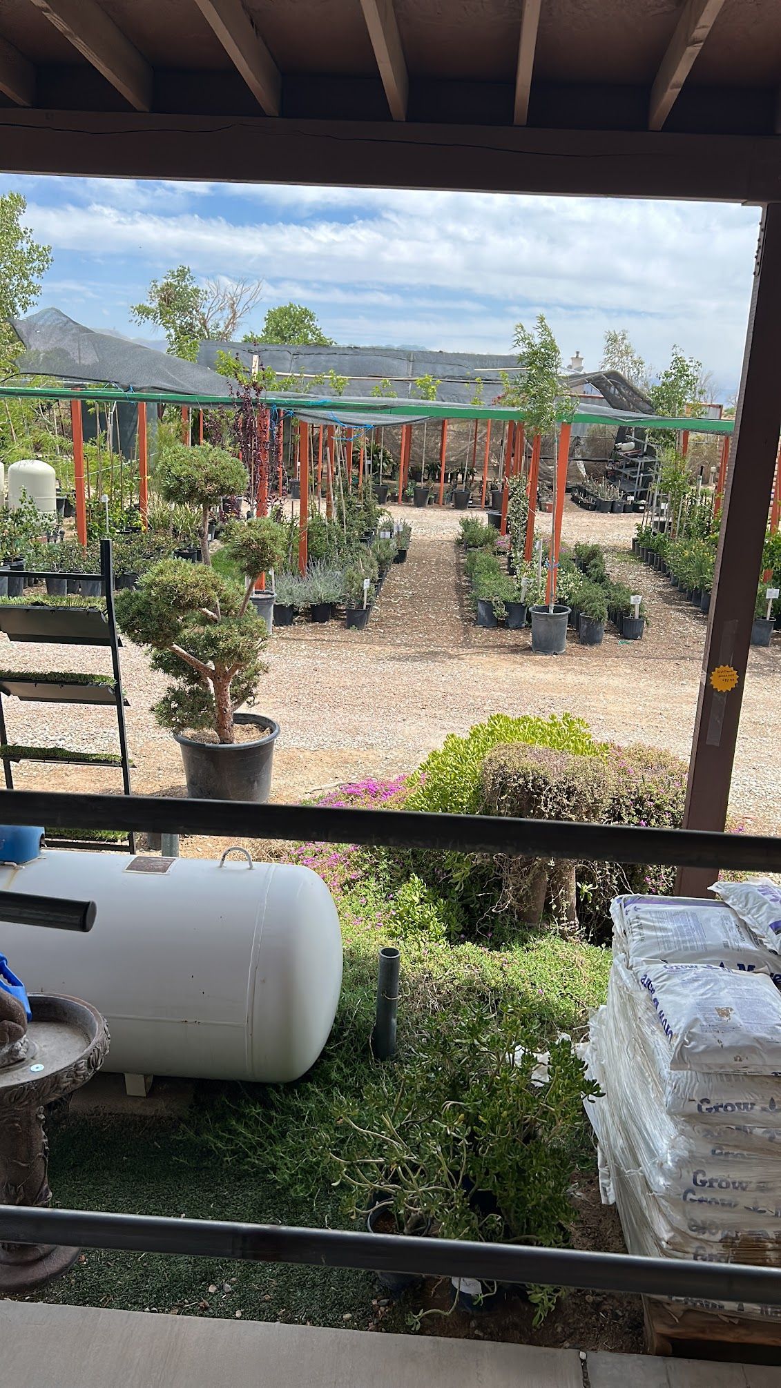 A plant nursery with rows of potted plants under shaded structures, view from a porch.