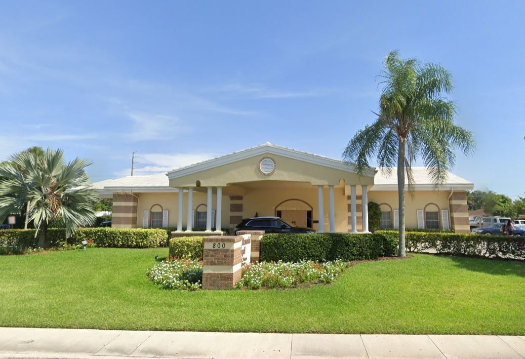 Yellow building with columns, green lawn, palm trees, and a brick mailbox.