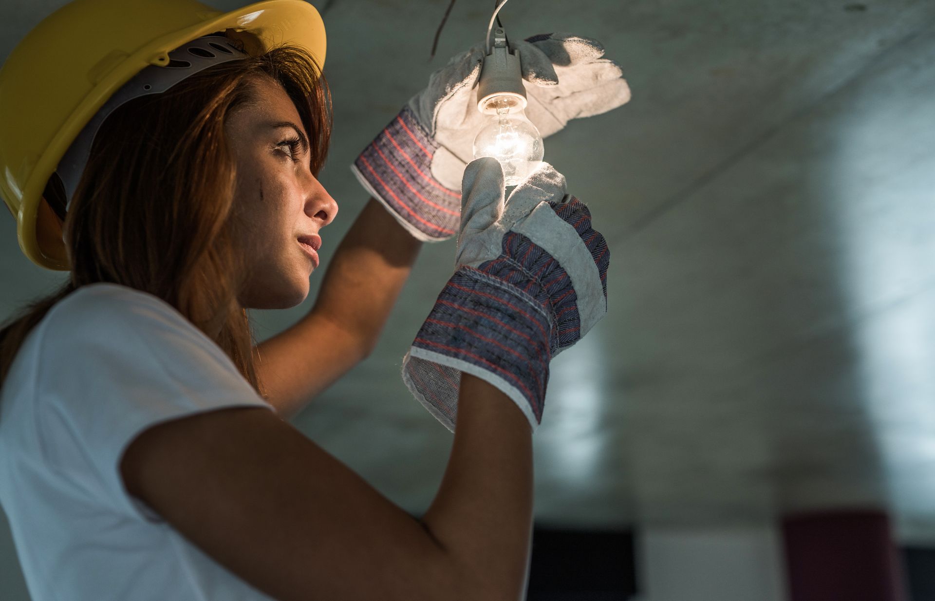 A woman in a hard hat and gloves is looking at a light bulb — Las Vegas, NV — Discount Lighting Supply